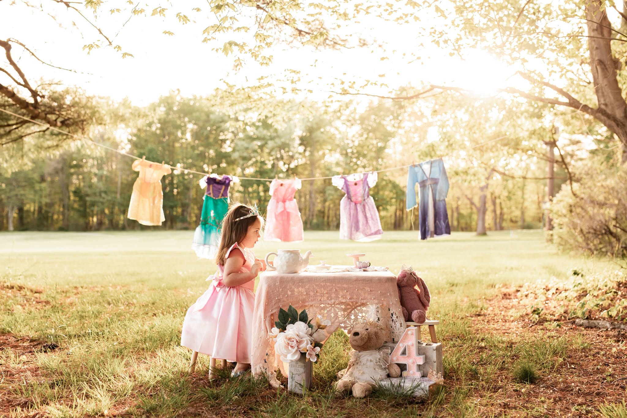 Girl in pink dress near stone fountain with stuffed animals, clothes hanging on line in park during sunset.