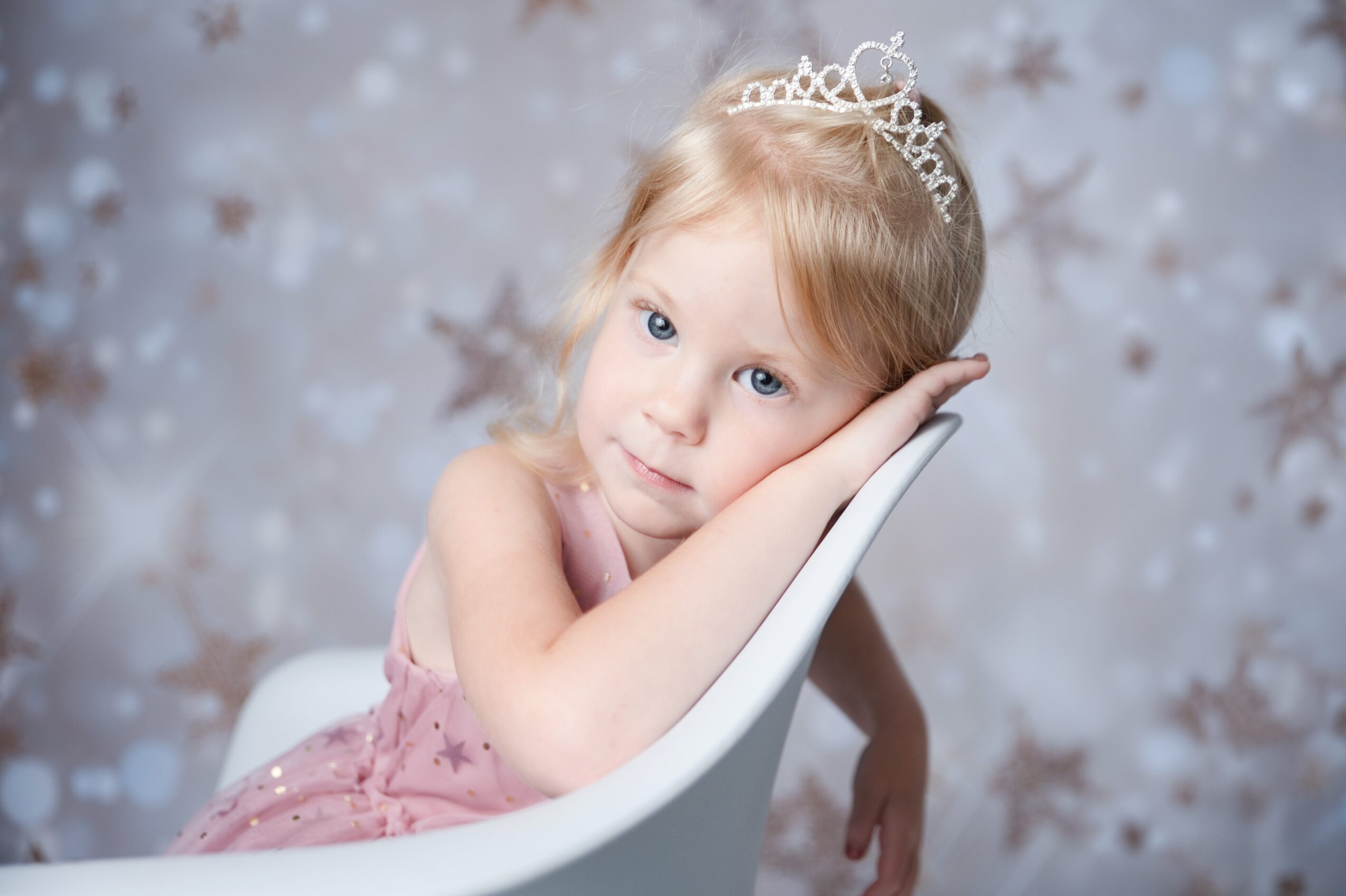 Young girl with blonde hair wearing a tiara, resting her head on her hand, sitting in a white chair, background with star patterns.