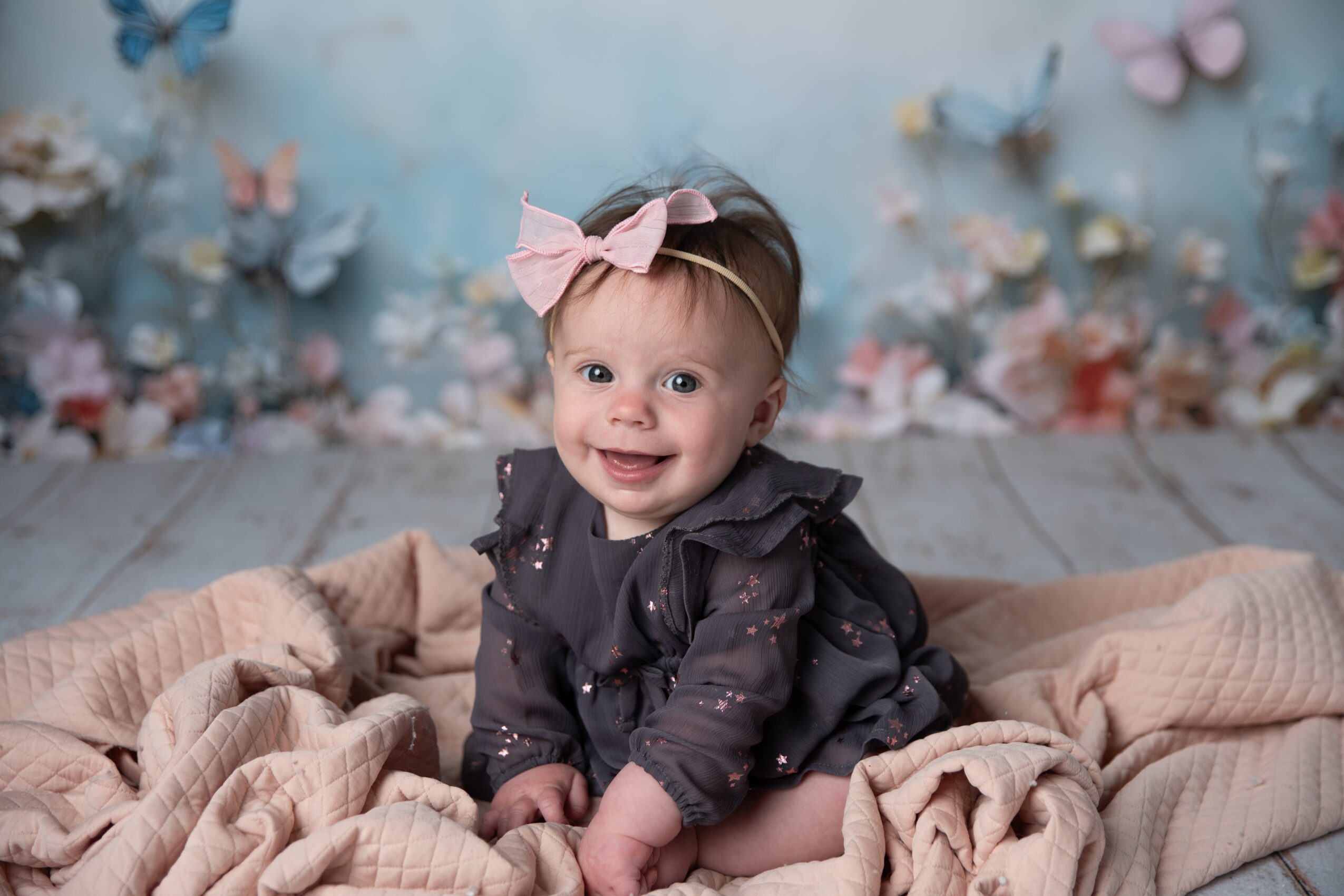 Smiling baby girl with a pink bow in her hair, sitting on a blanket with a floral background.