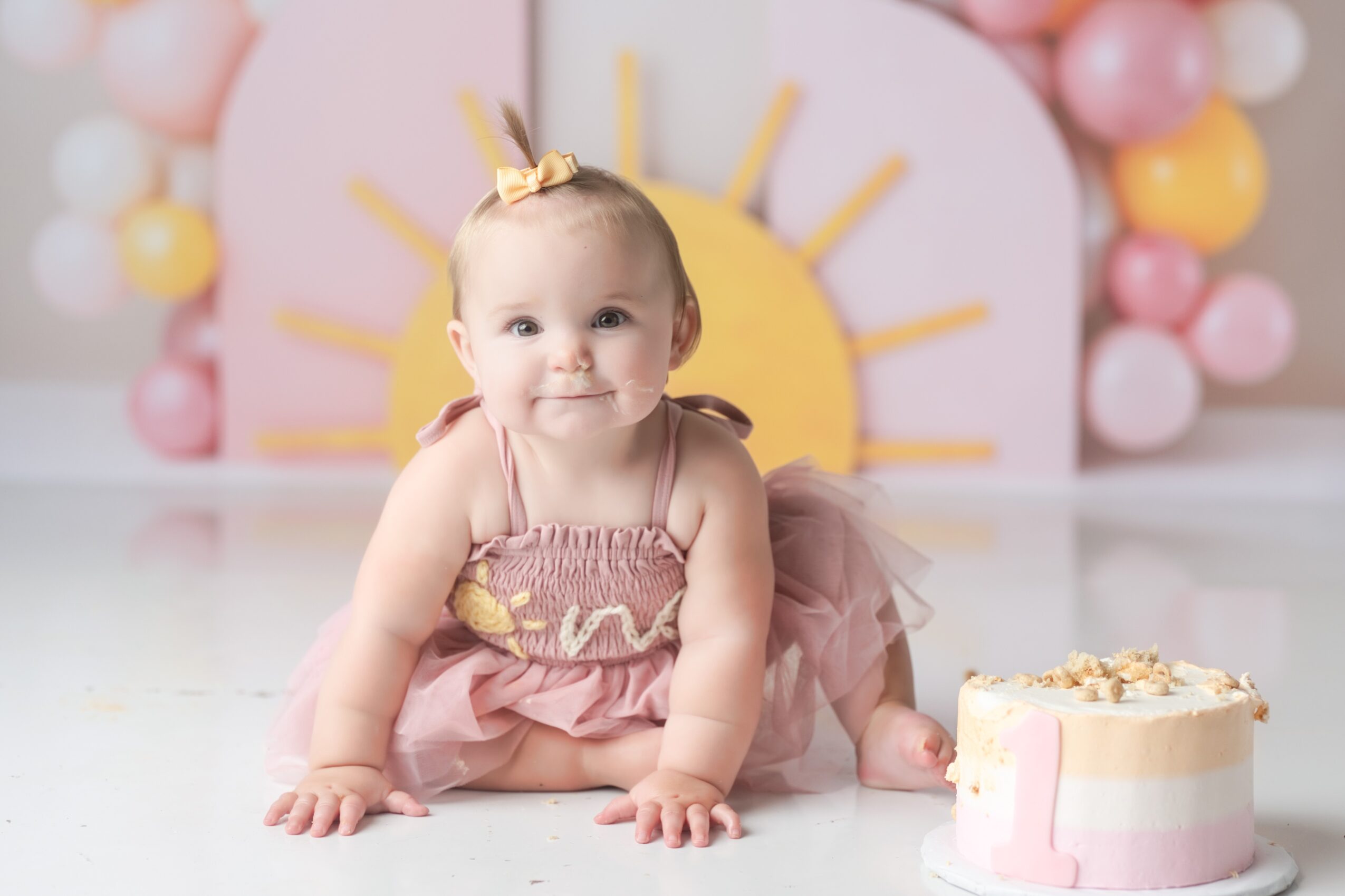 Baby girl in pink dress crawling on floor with birthday cake nearby, colorful balloons and sun decoration in background.