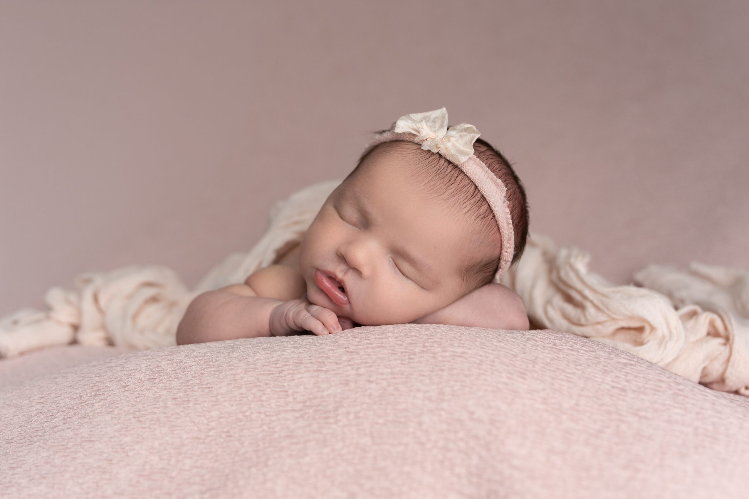 Sleeping baby with a headband resting on a soft surface, with a blanket behind her.