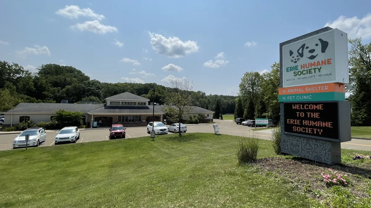 Building with parking lot and cars, sign for Humane Society, grassy area, trees, blue sky with clouds.
