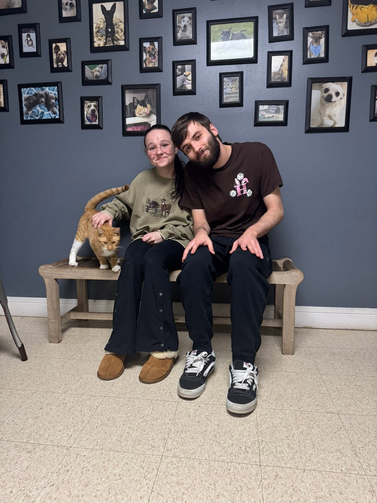 Two people sitting on a wooden bench in front of a wall with framed photos, with a cat beside them.
