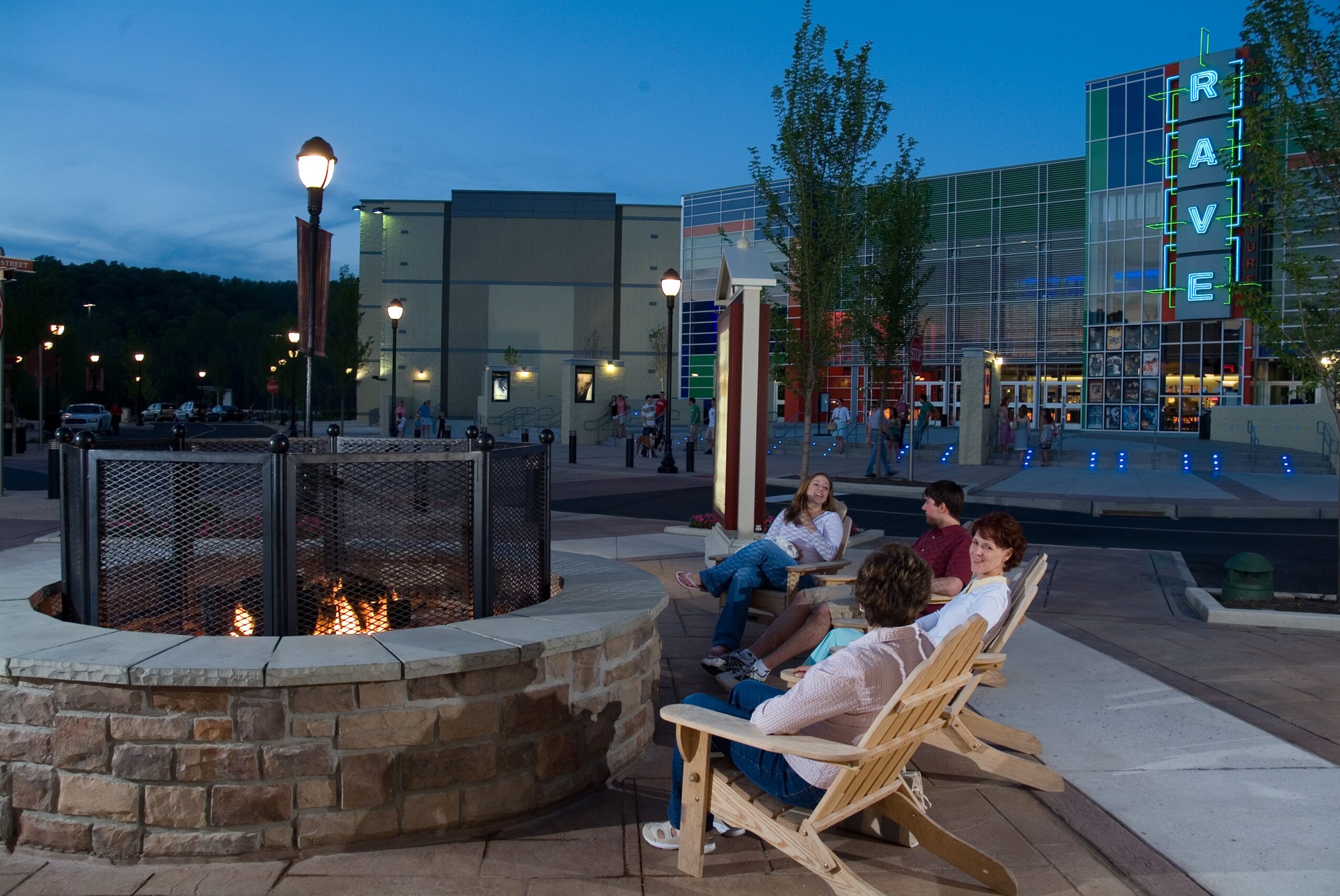 People sit on chairs near a fire pit outside a building with a neon sign reading 'RAVE' during evening.