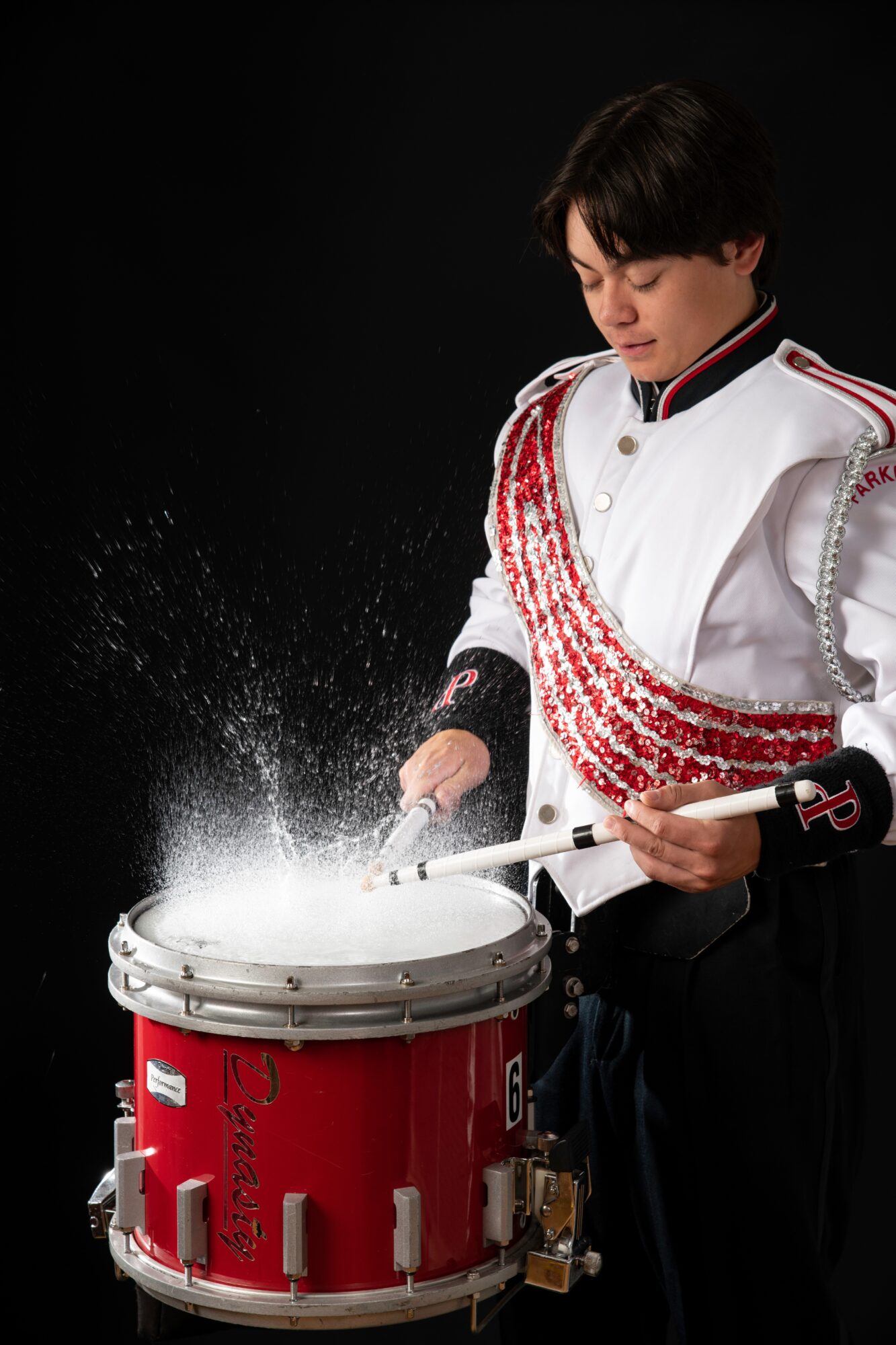A young person in a white and red marching band uniform plays a snare drum with drumsticks, with water splashing.