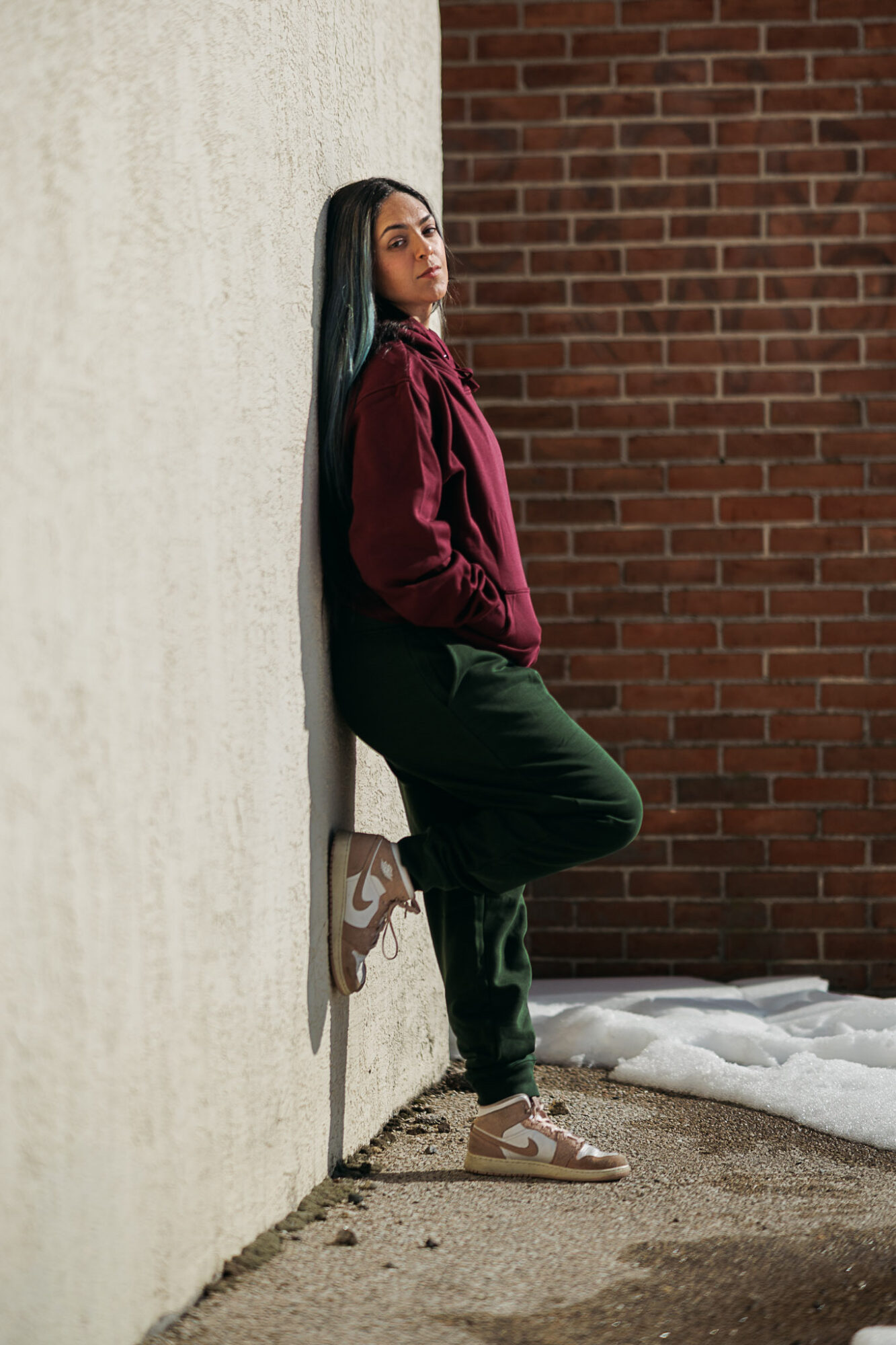 Person leaning against a wall outdoors, wearing a maroon jacket and dark pants, with snow on the ground.