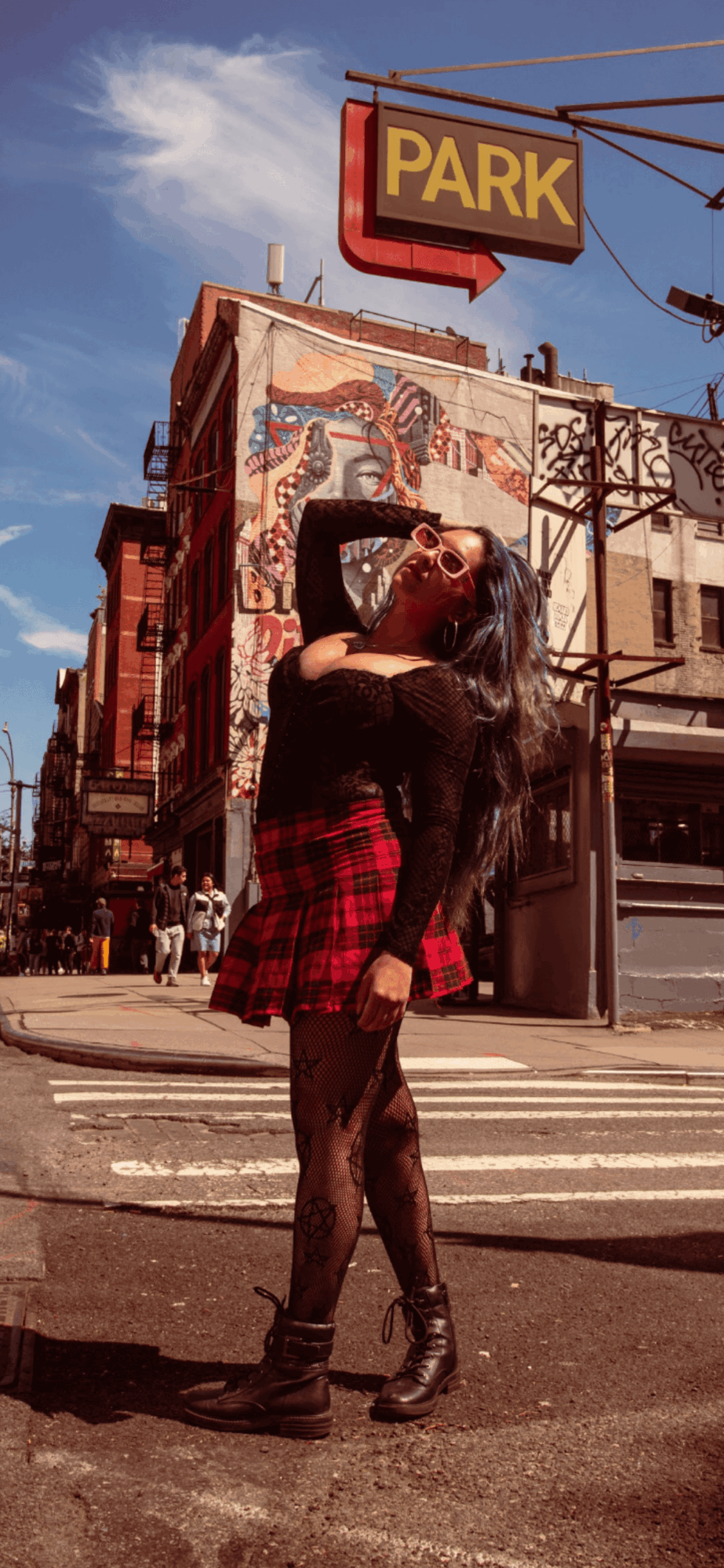 Person with long dark hair wearing a black top and red plaid skirt standing on a city street corner.
