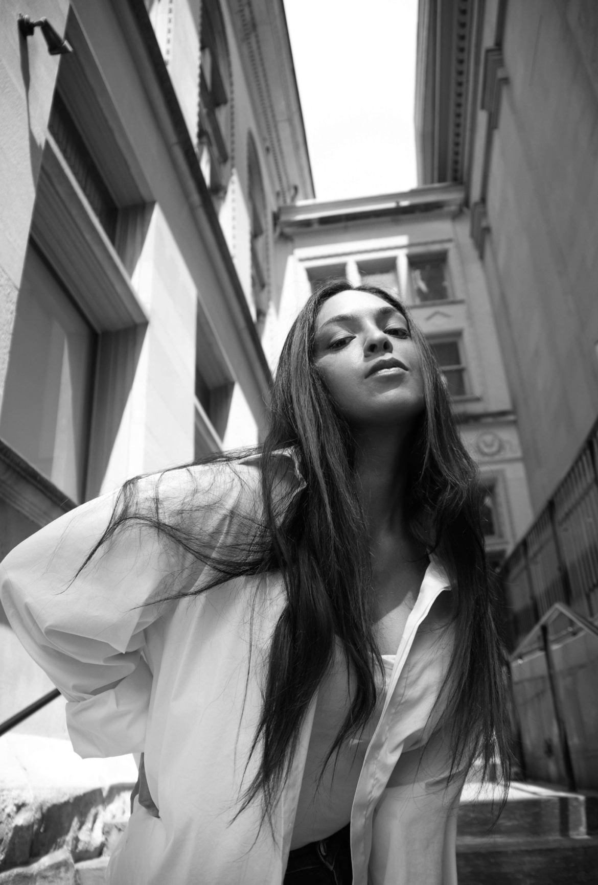 Young woman with long hair in a white shirt standing outdoors between tall buildings, looking at the camera.