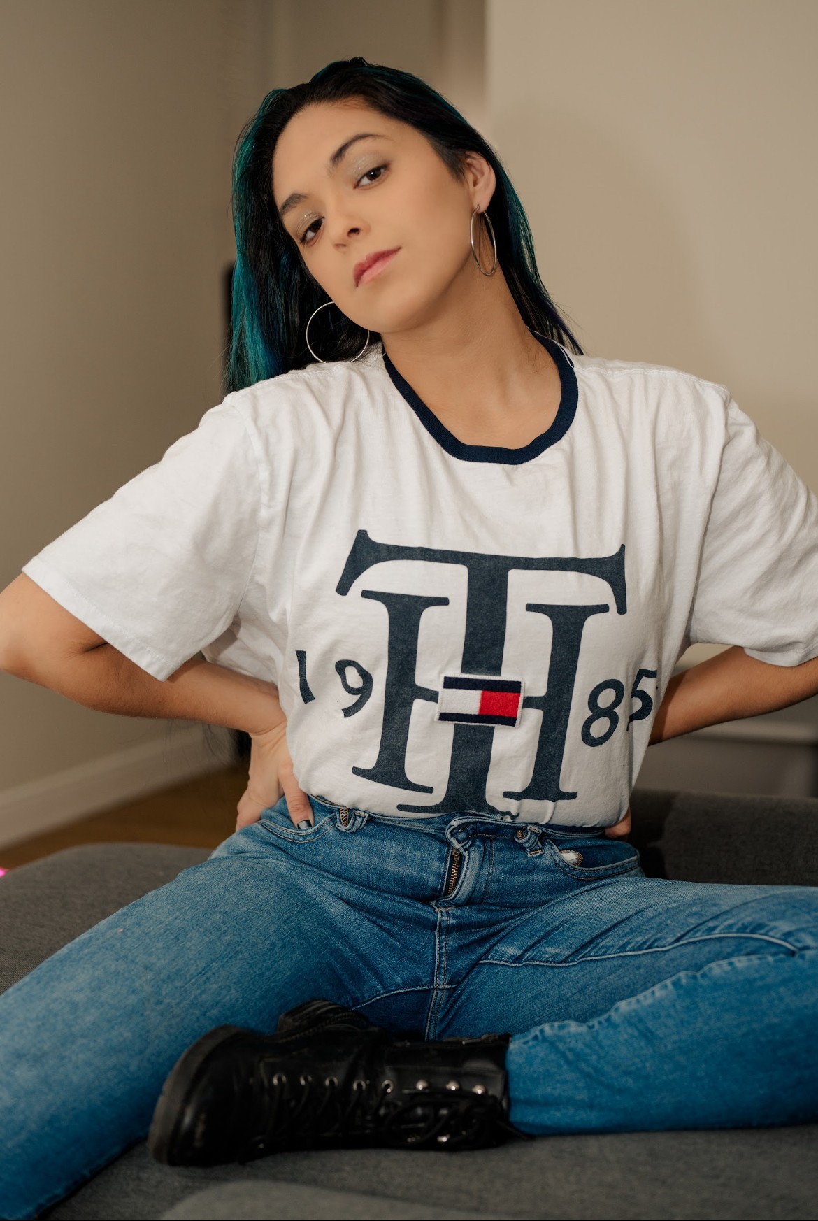 Young woman with dark hair and hoop earrings, wearing a white T-shirt and jeans, sitting on a couch with hands on hips.