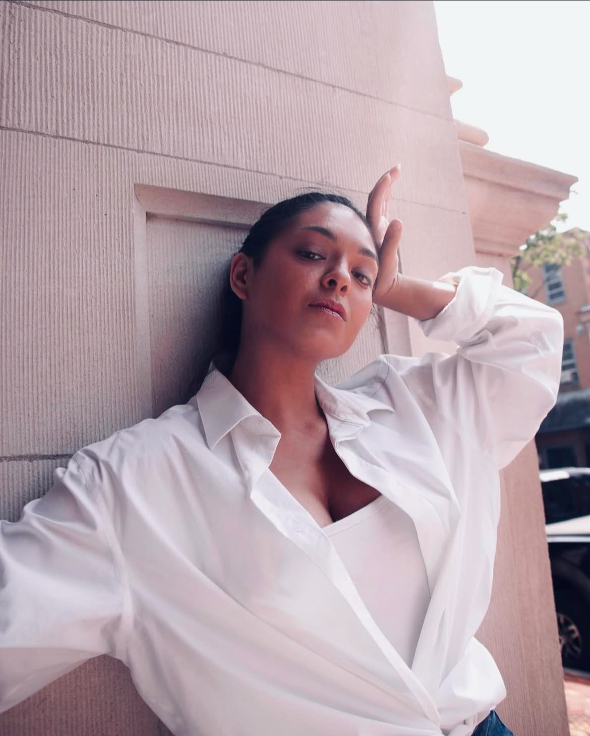 Woman with dark hair in a white shirt, posing with hand on forehead, leaning against a wall outdoors.