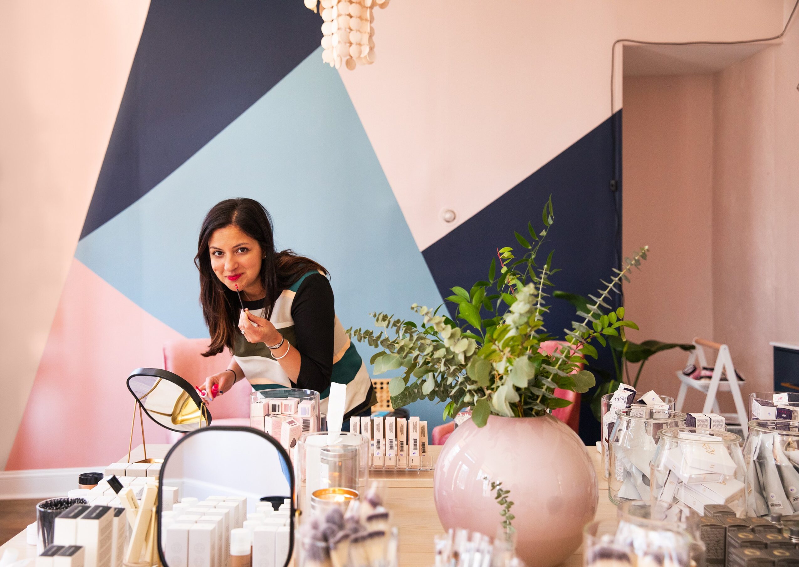 Woman with dark hair holding a cup, standing behind a table with various items, in front of a geometric wall design.
