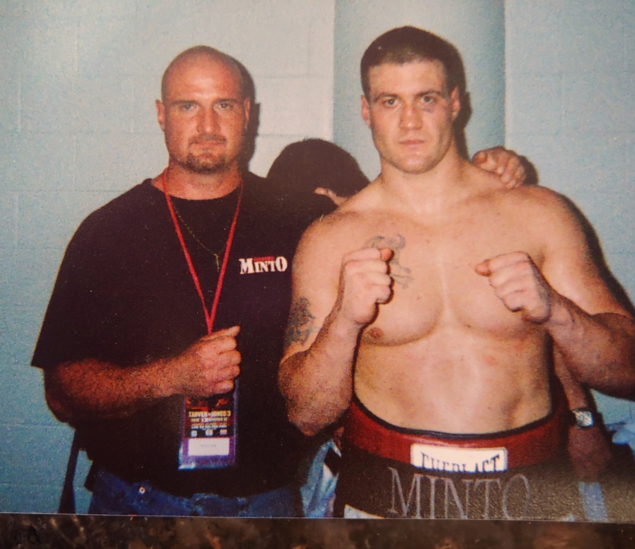 Two men, one shirtless with fists raised, the other in a black shirt, standing against a light blue wall.