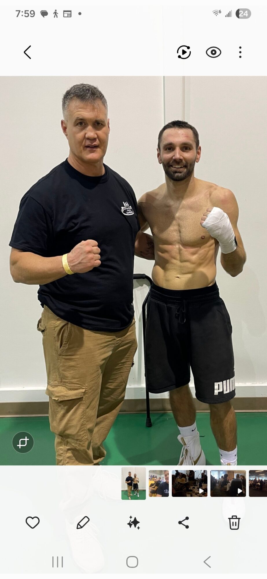 Two men standing side by side, one with a fist raised, the other shirtless with boxing gloves, smiling.