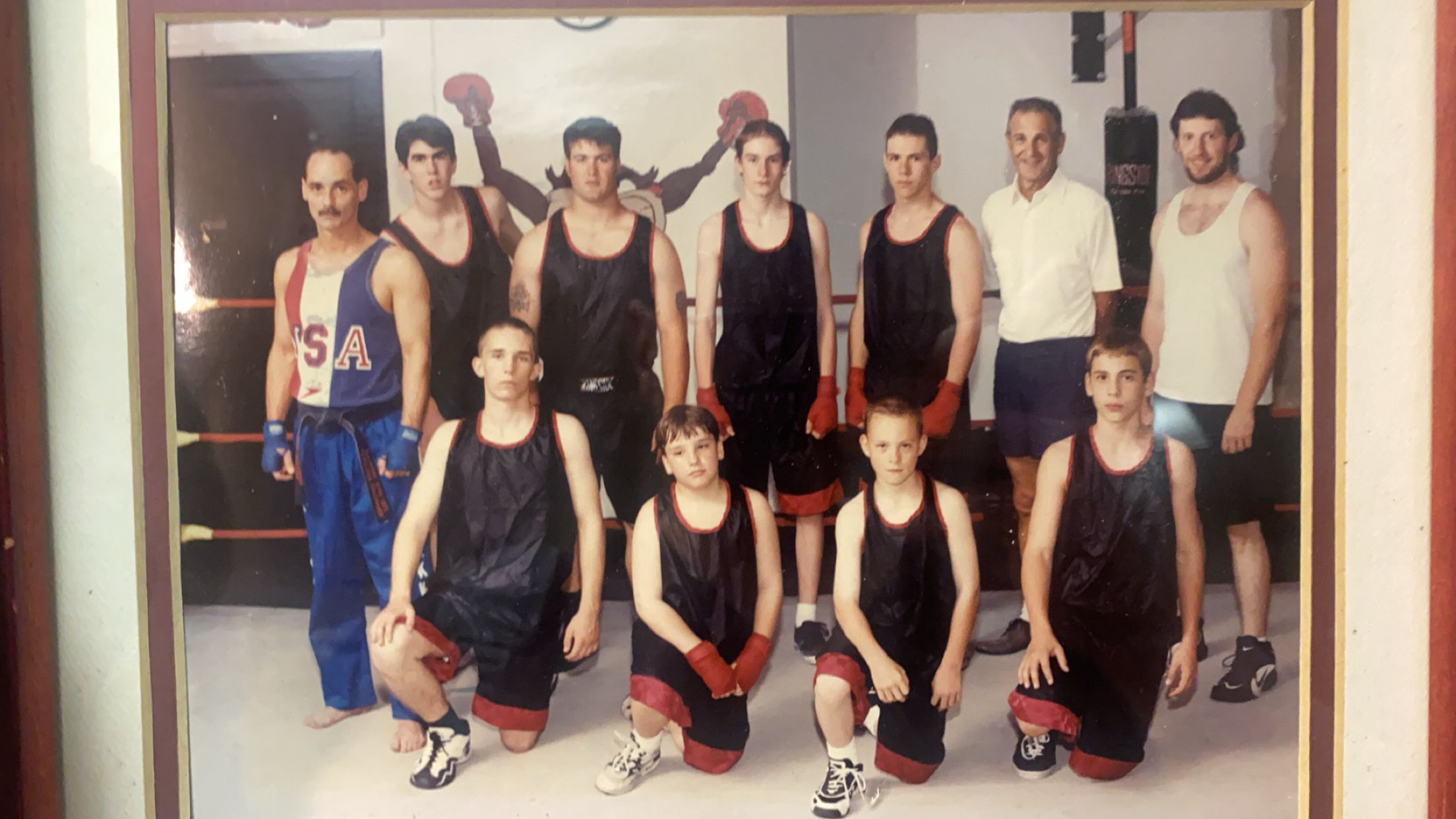 Group of ten people in athletic clothing standing indoors, some with hands on hips, in front of a mirror or window.