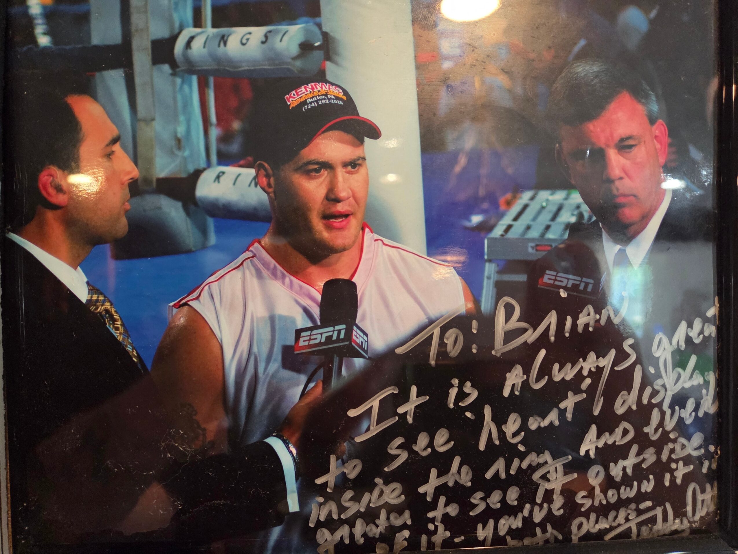 Three men, one in a sports jersey, speaking into microphones, with a boxing ring in the background.