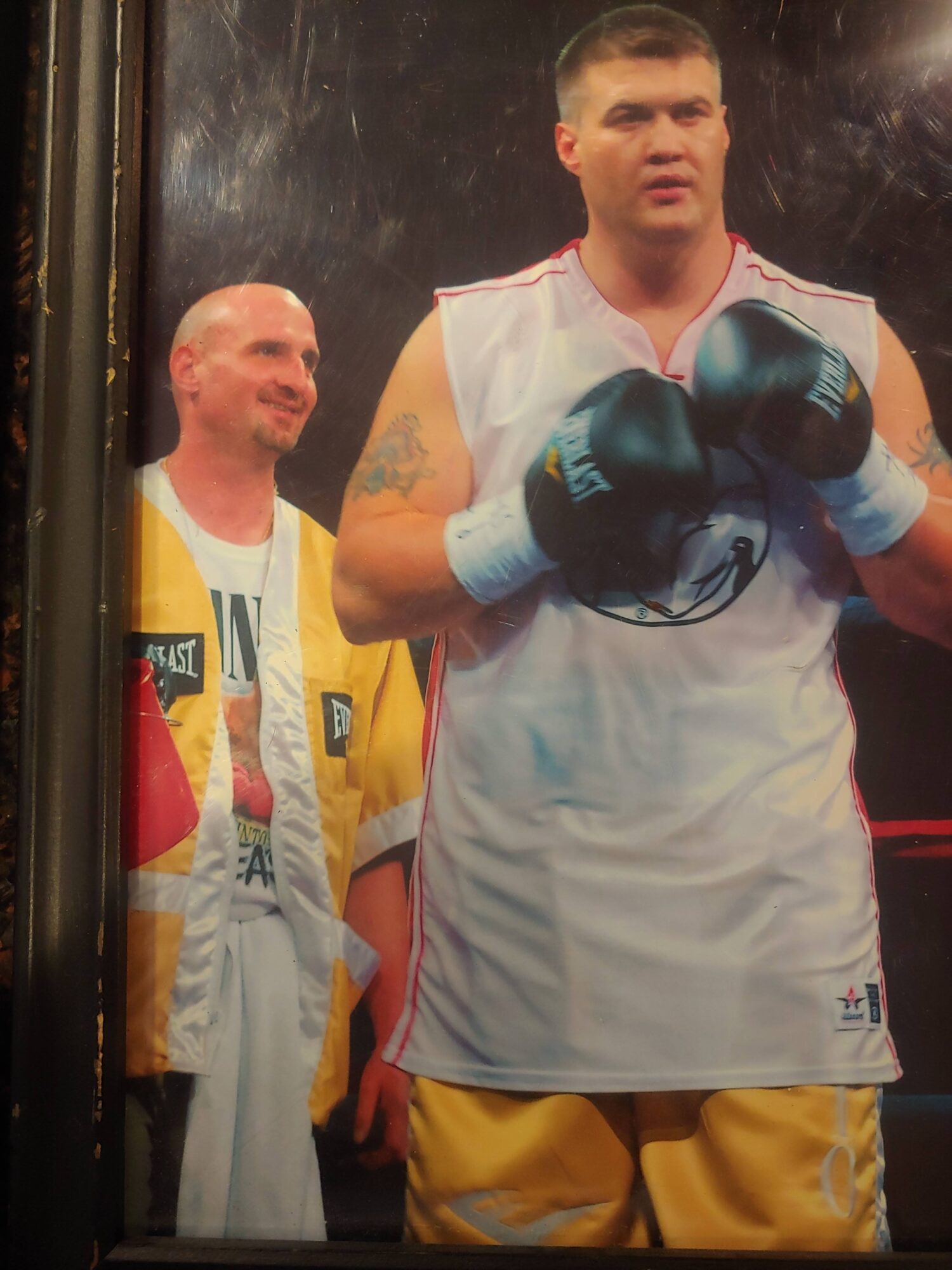 Two young men in boxing gloves and athletic clothing, one with a tattoo, standing in a gym or training area.