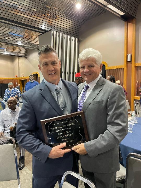 Two men in suits holding a plaque at a formal event, smiling, with other people seated at tables in the background.