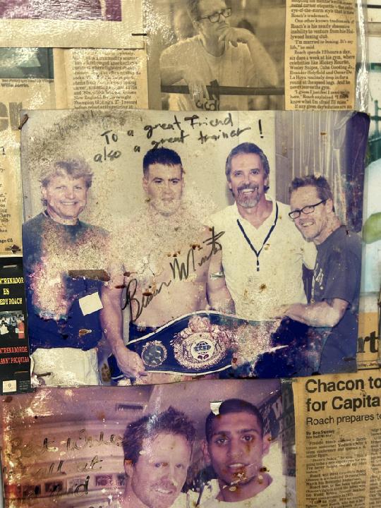 Four men standing together, one with a championship belt, smiling, with handwritten note above. Background includes newspaper clippings.