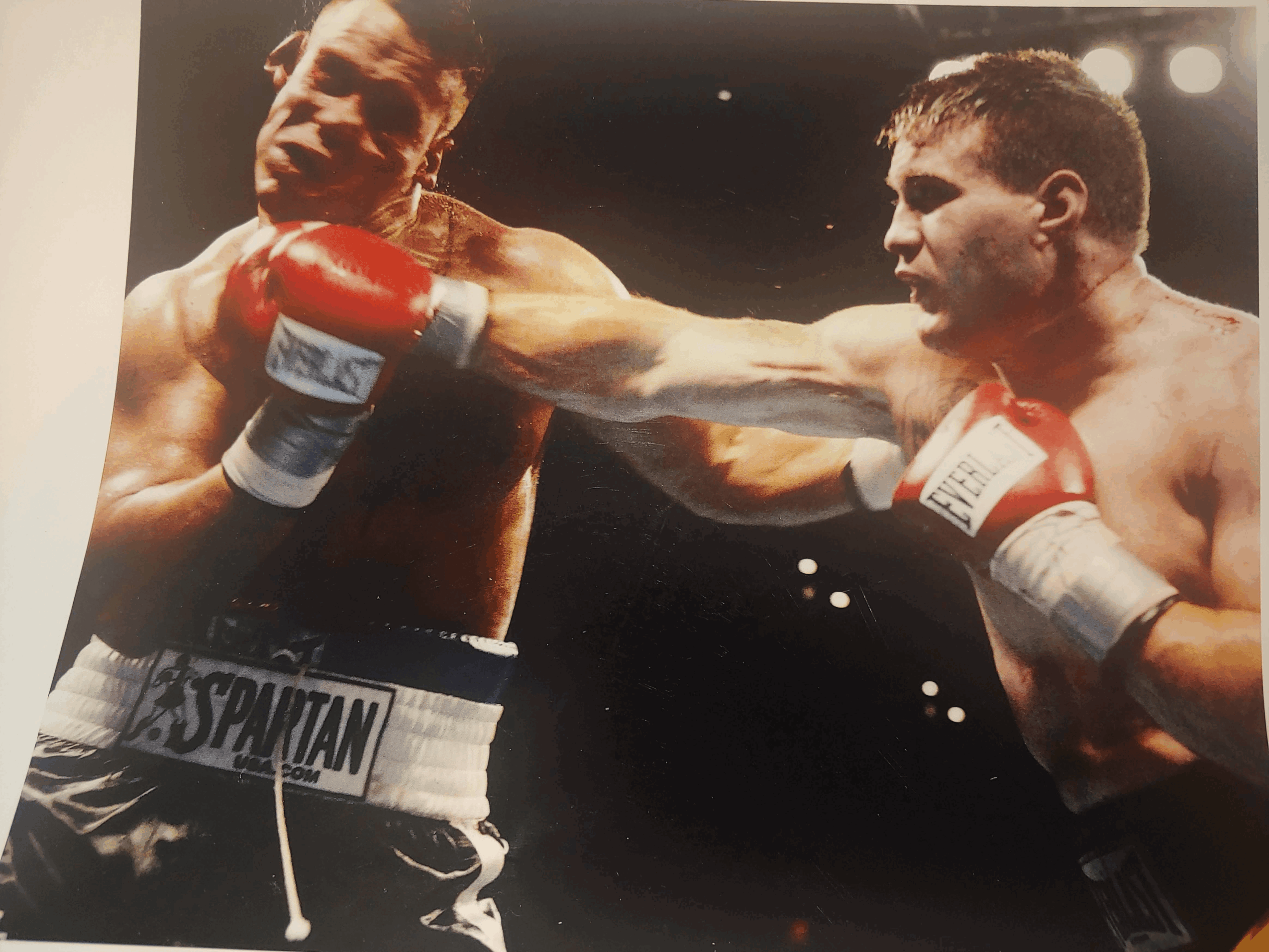 Two boxers in a match, one delivering a punch to the other's face, both wearing boxing gloves and shorts.