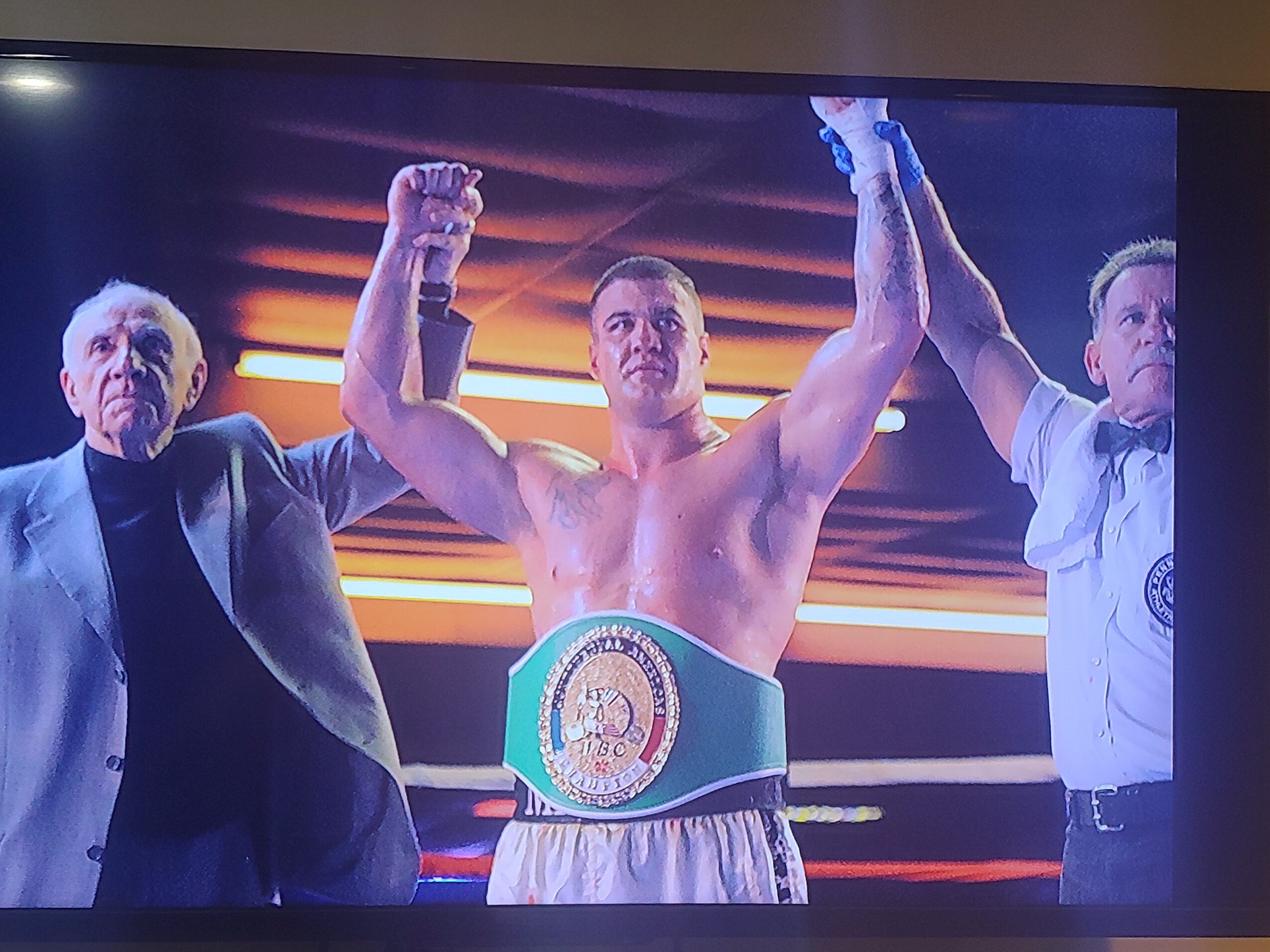 Boxer with arms raised, wearing a championship belt, flanked by two officials, in a boxing ring with bright lights.