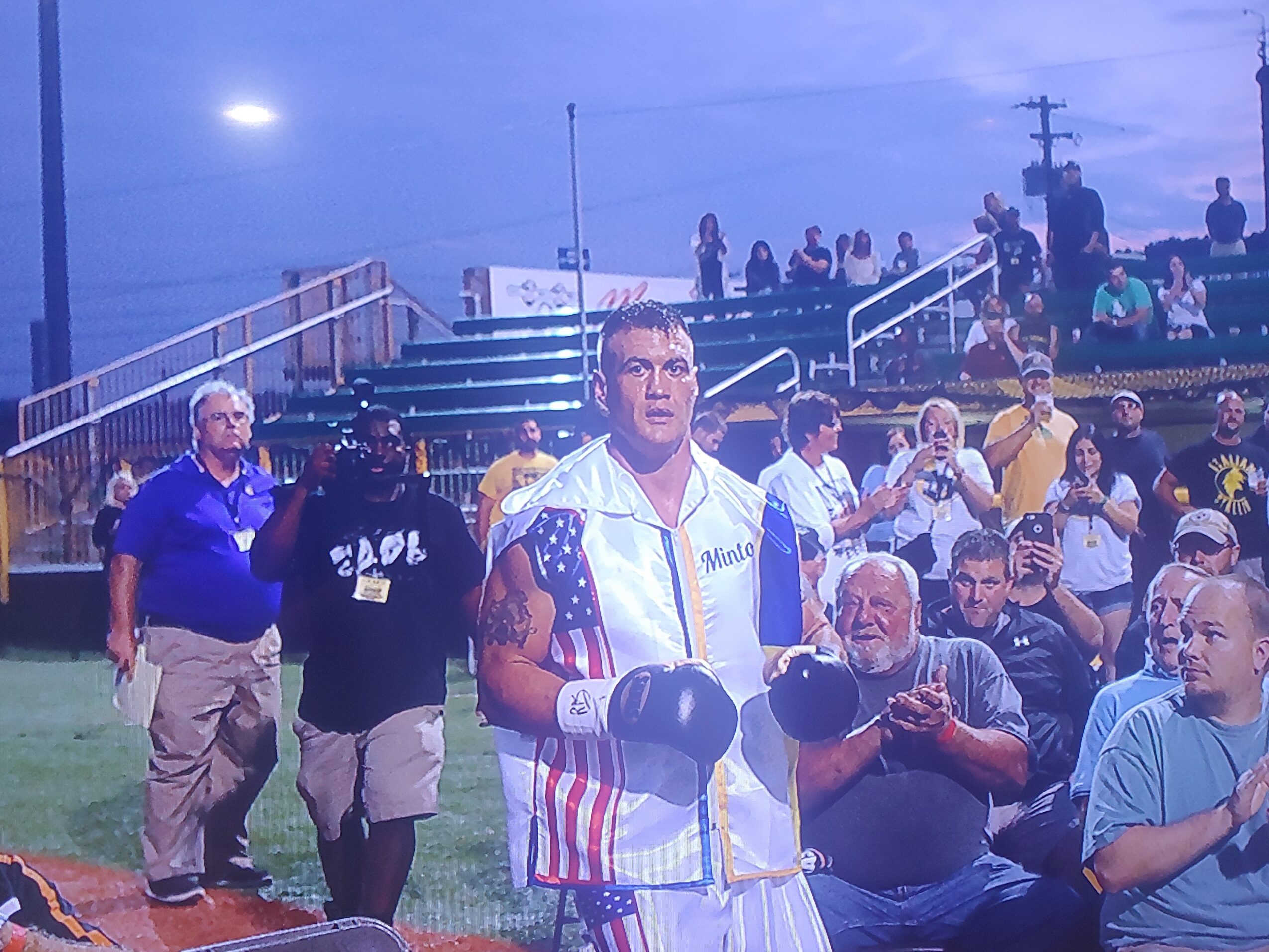 Boxer in white and blue robe with gloves stands in a crowded outdoor area with spectators and staff, evening sky visible.