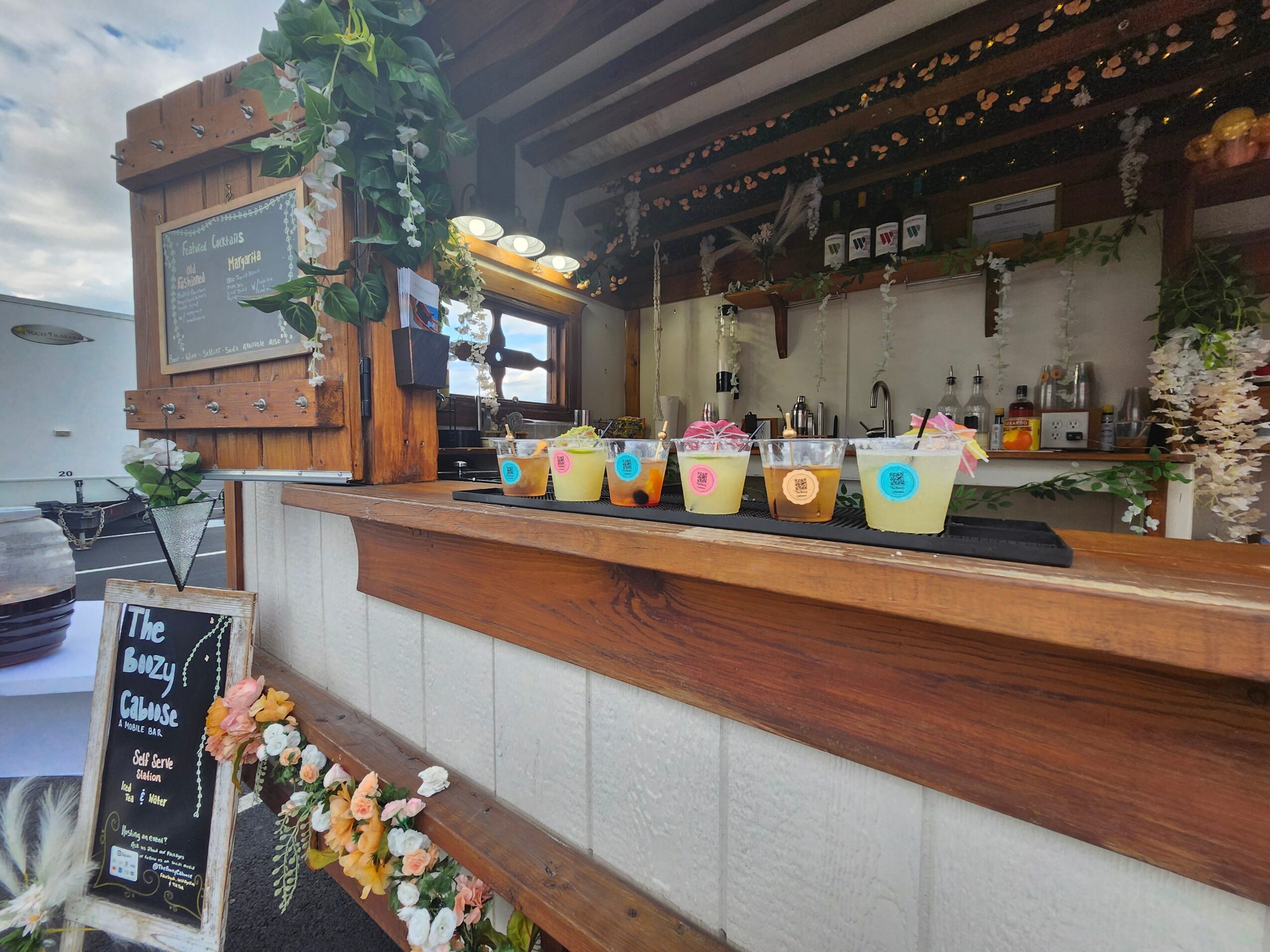 Bar with colorful drinks in cups, wooden counter, and decorative flowers, with a chalkboard menu and plants in the background.