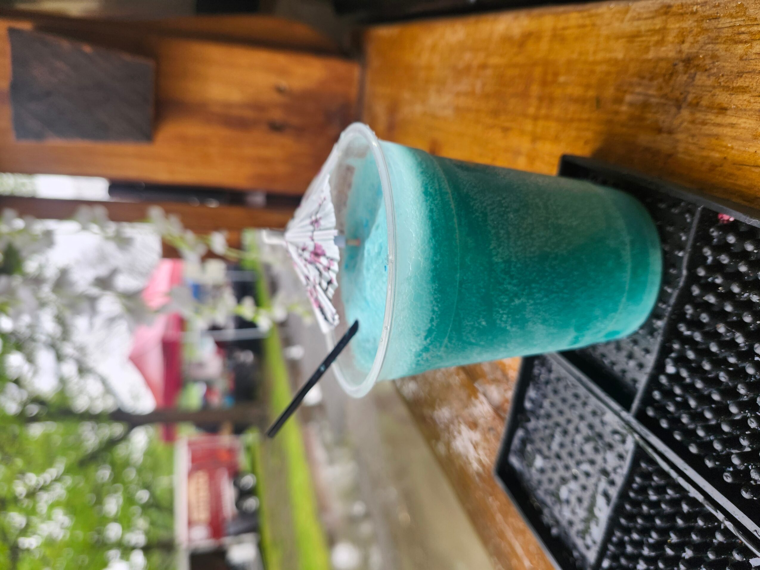 Cold blue beverage in a clear plastic cup with a black straw on a wooden table outdoors.
