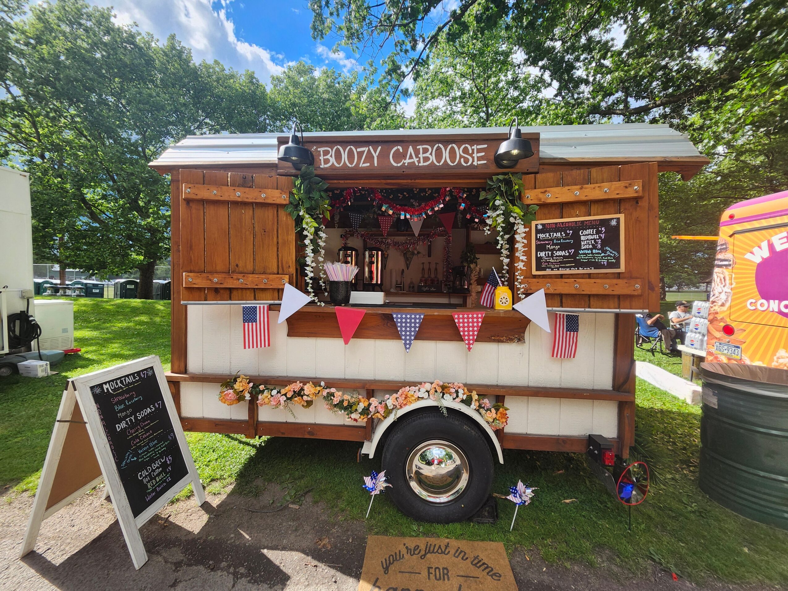 Wooden food truck decorated with banners, flowers, and a sign reading 'Boozy Caboose' in a park setting.