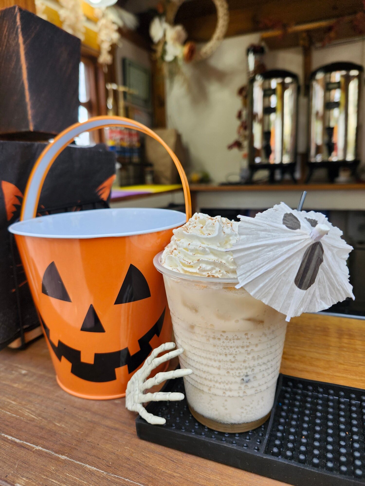 A Halloween-themed drink in a pumpkin-shaped container with a black lid, decorated with a white paper napkin, a straw, and a small white skeleton hand, on a wooden table.
