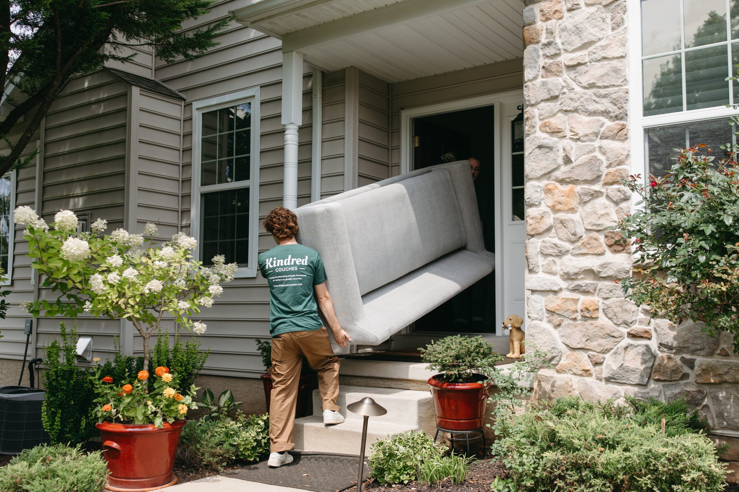 Person moving a sofa into a house through a front door, surrounded by plants and flowers.