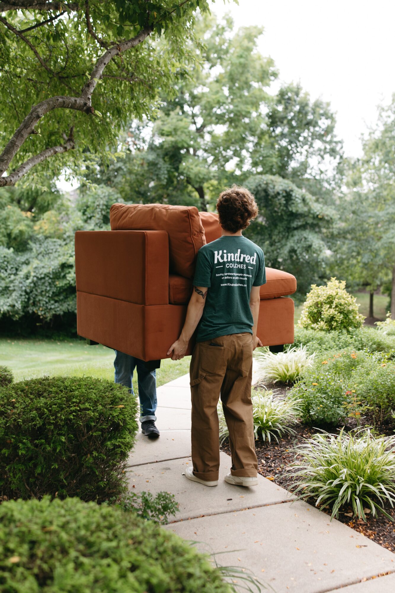 Two people carry a brown sofa along a garden path surrounded by greenery and plants.