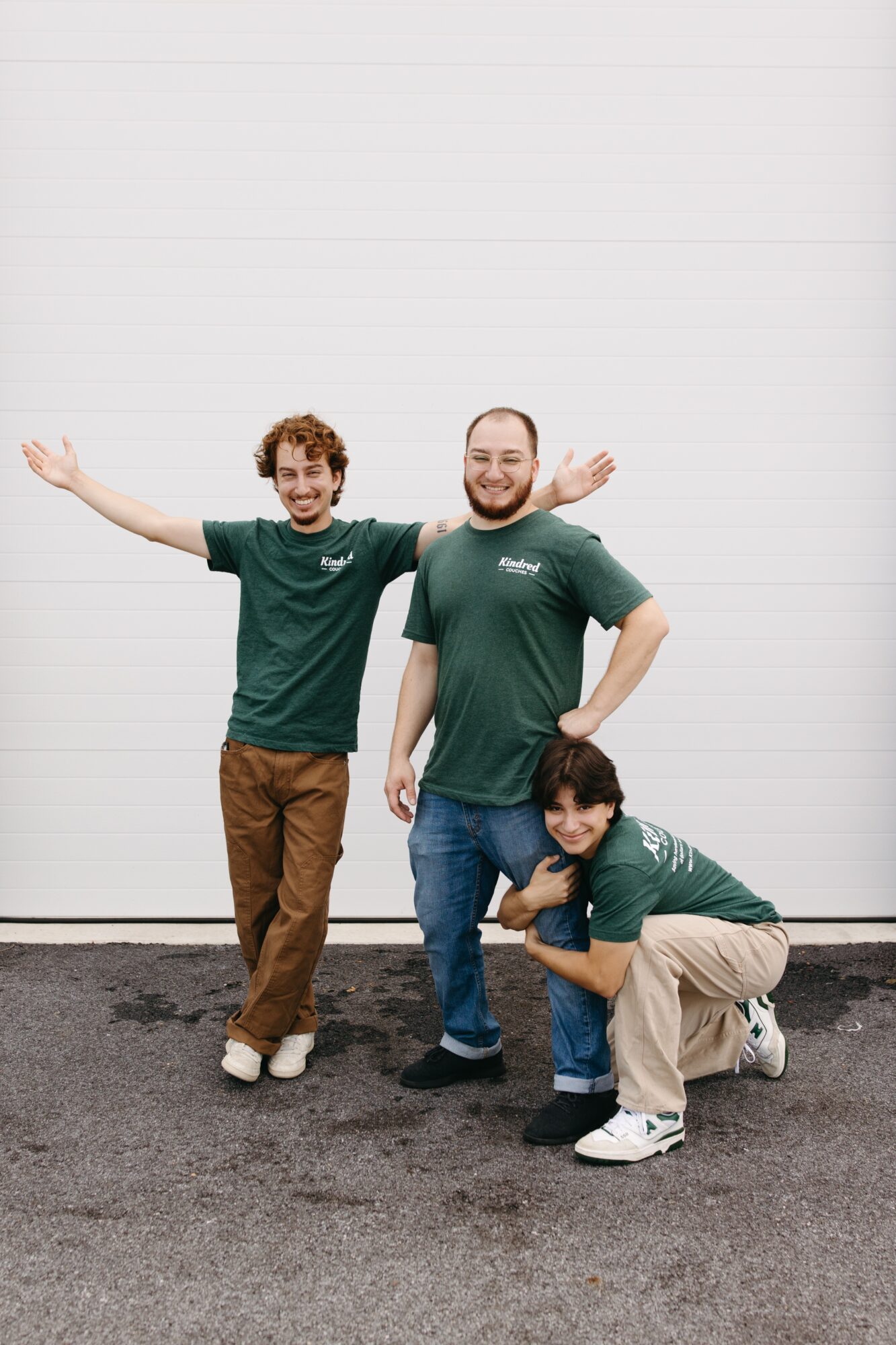 Three people in green shirts standing and crouching outdoors against a white wall, smiling and posing with arms outstretched.
