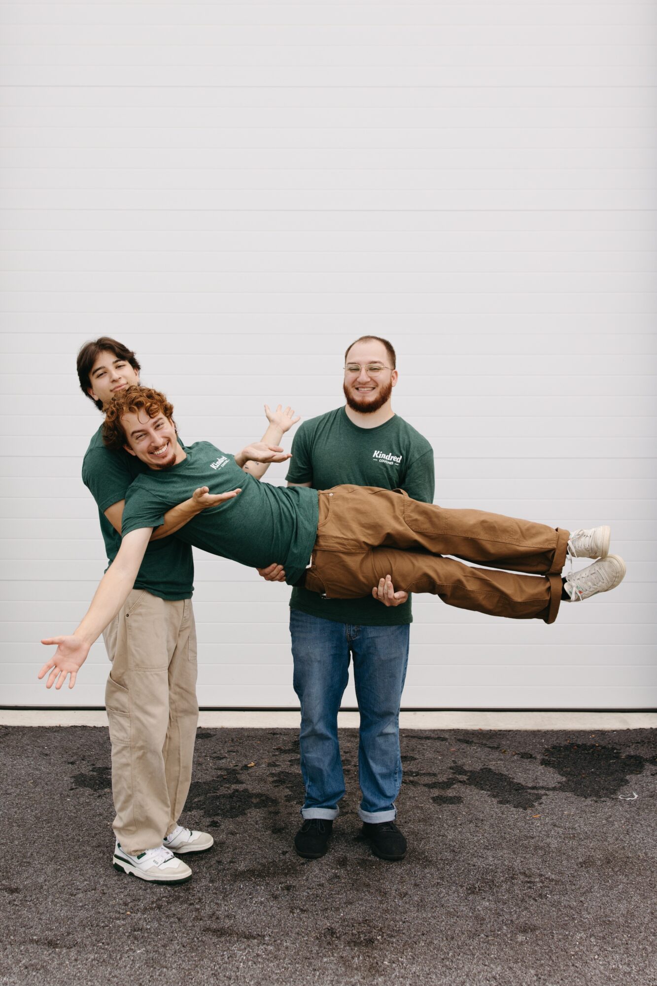 Three people standing outside, two holding a person horizontally, all smiling, against a white wall background.