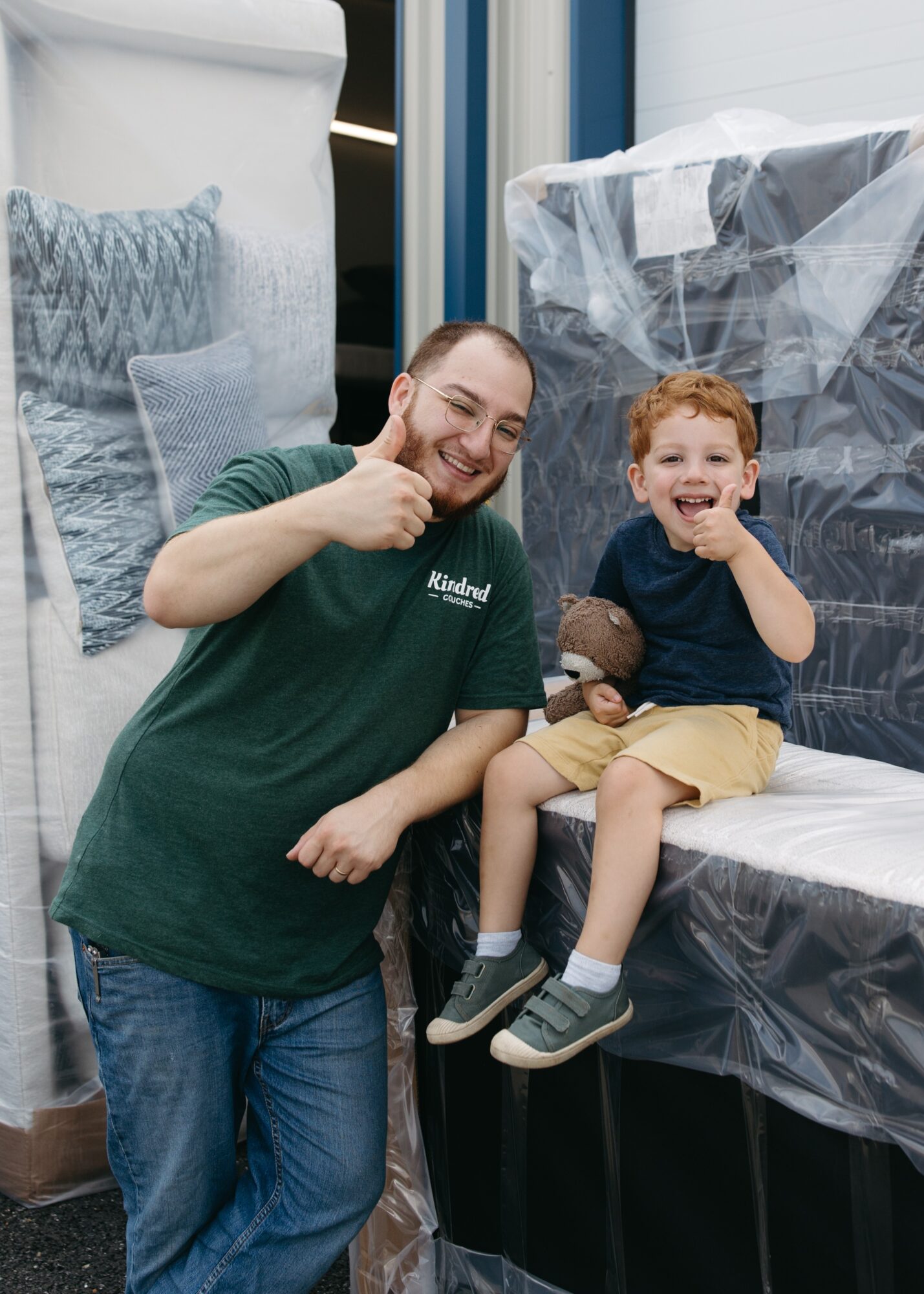 Two people, a man and a young boy, sit on a hospital bed, smiling and giving thumbs up, with medical equipment in background.