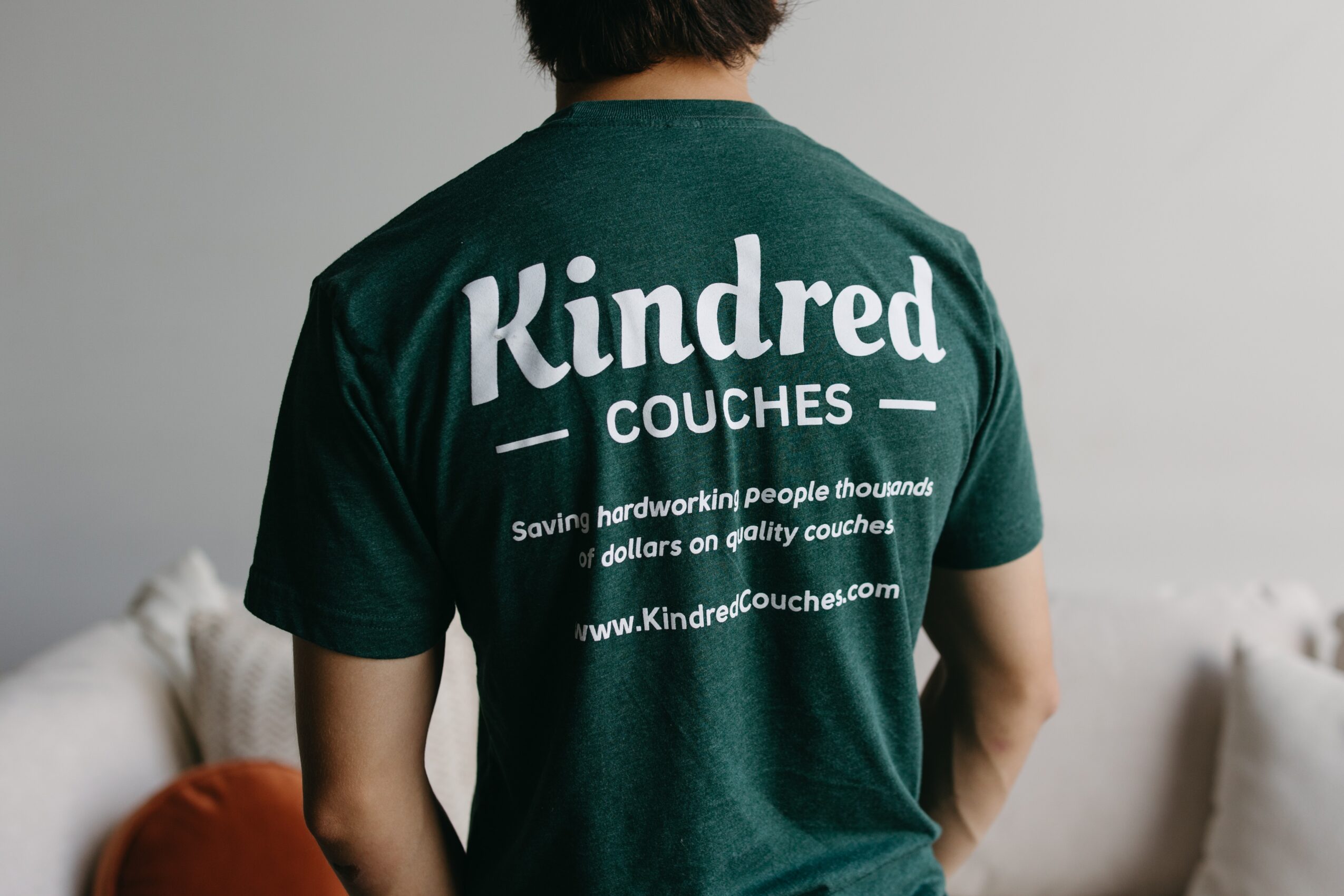 Person wearing a green T-shirt with white text on the back, sitting in a room with a sofa and cushions.