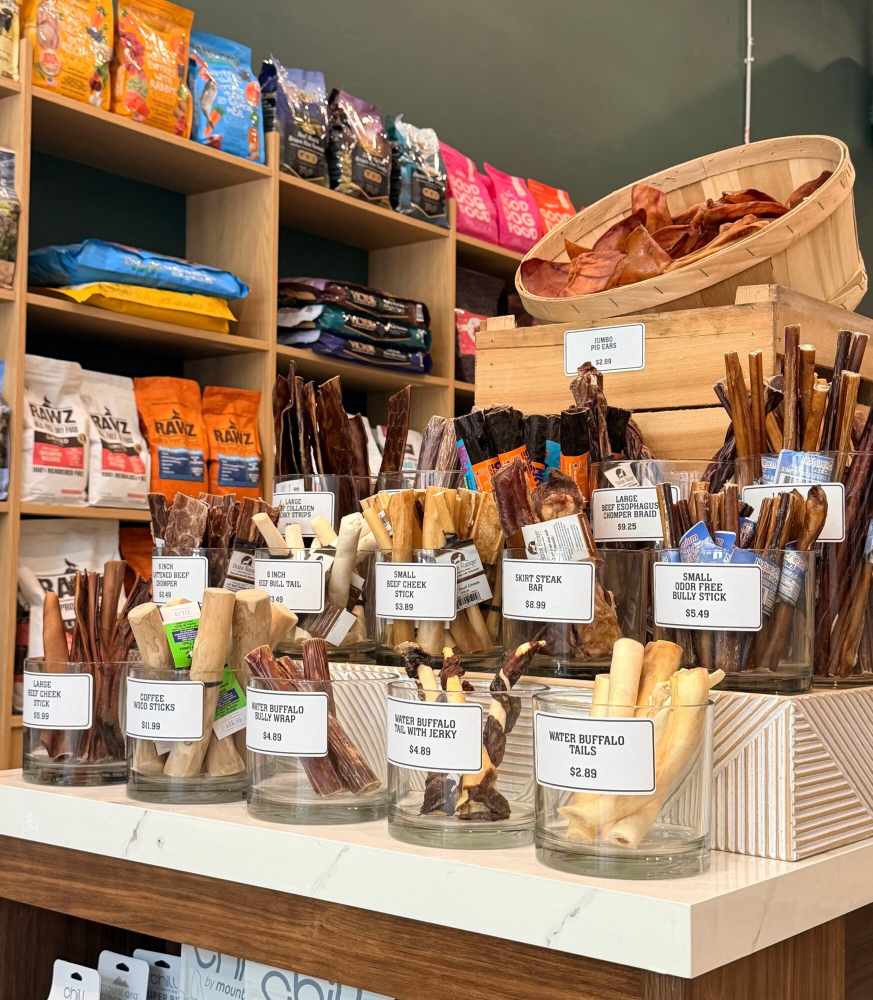 Display of various chocolate and snack products in glass jars with labels on a store counter, shelves with snack bags in background.