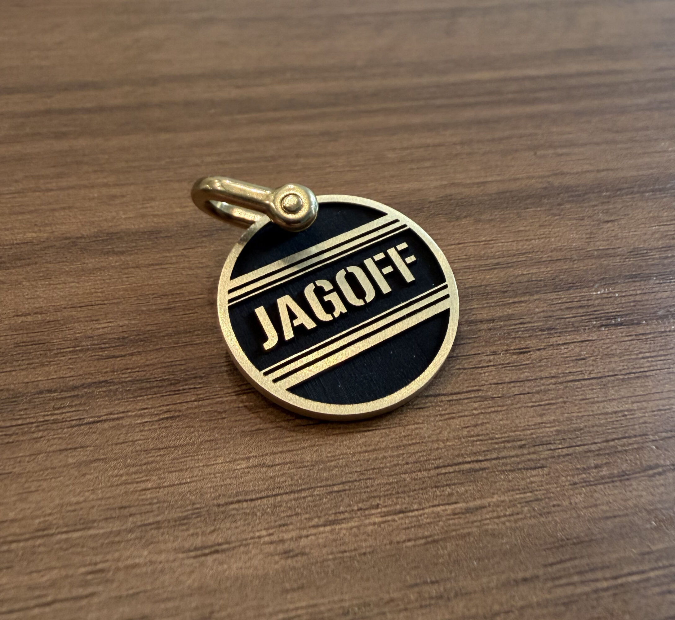 Round gold and black pin with the word JAGOFF and two horizontal lines, on a wooden surface.