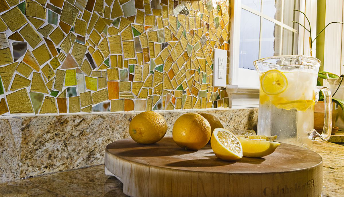 Lemon slices and whole lemons on a wooden cutting board in a kitchen, with a mosaic tile backsplash and a glass pitcher of lemon water.