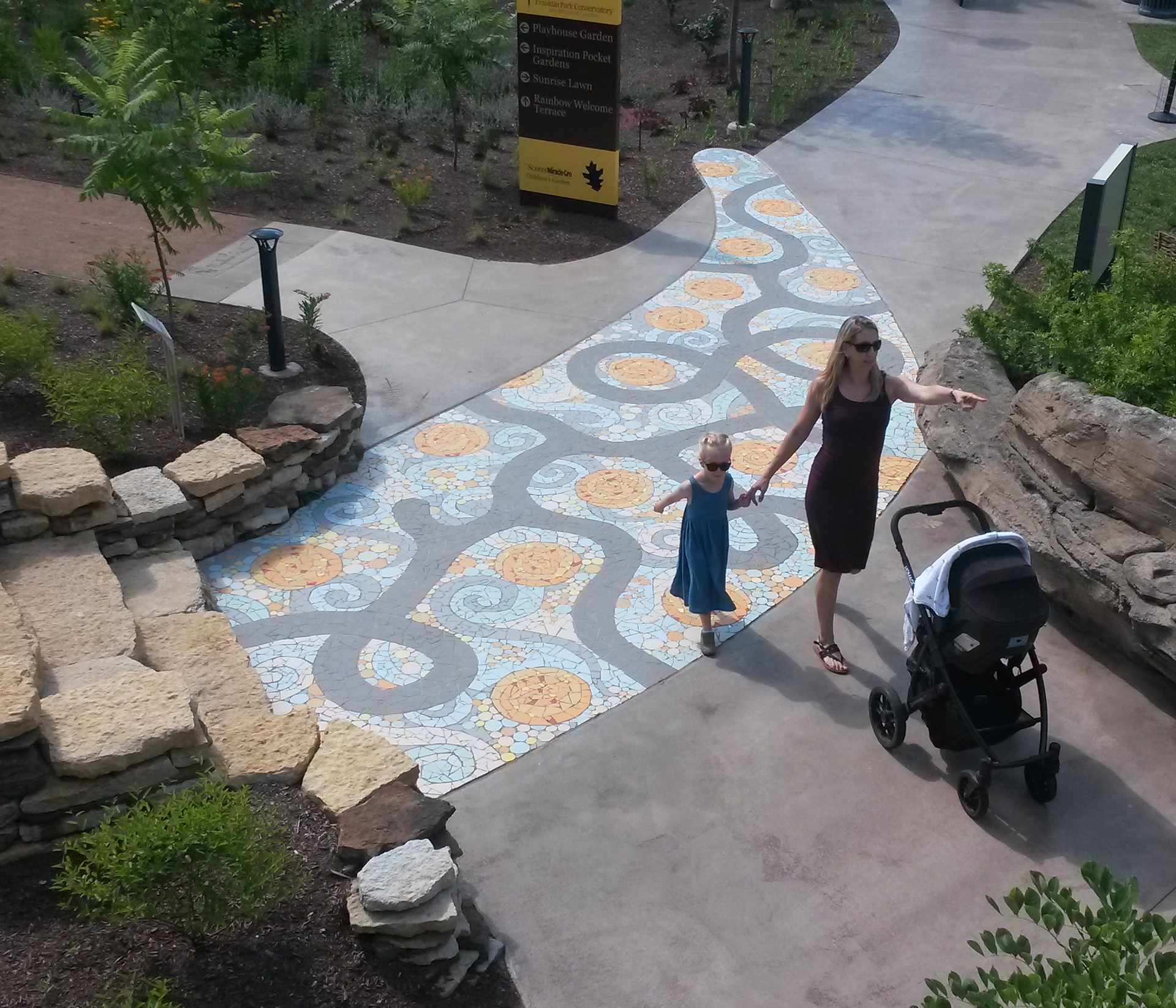 Woman and child walking on a colorful, winding pathway with a stroller nearby, surrounded by greenery and rocks.