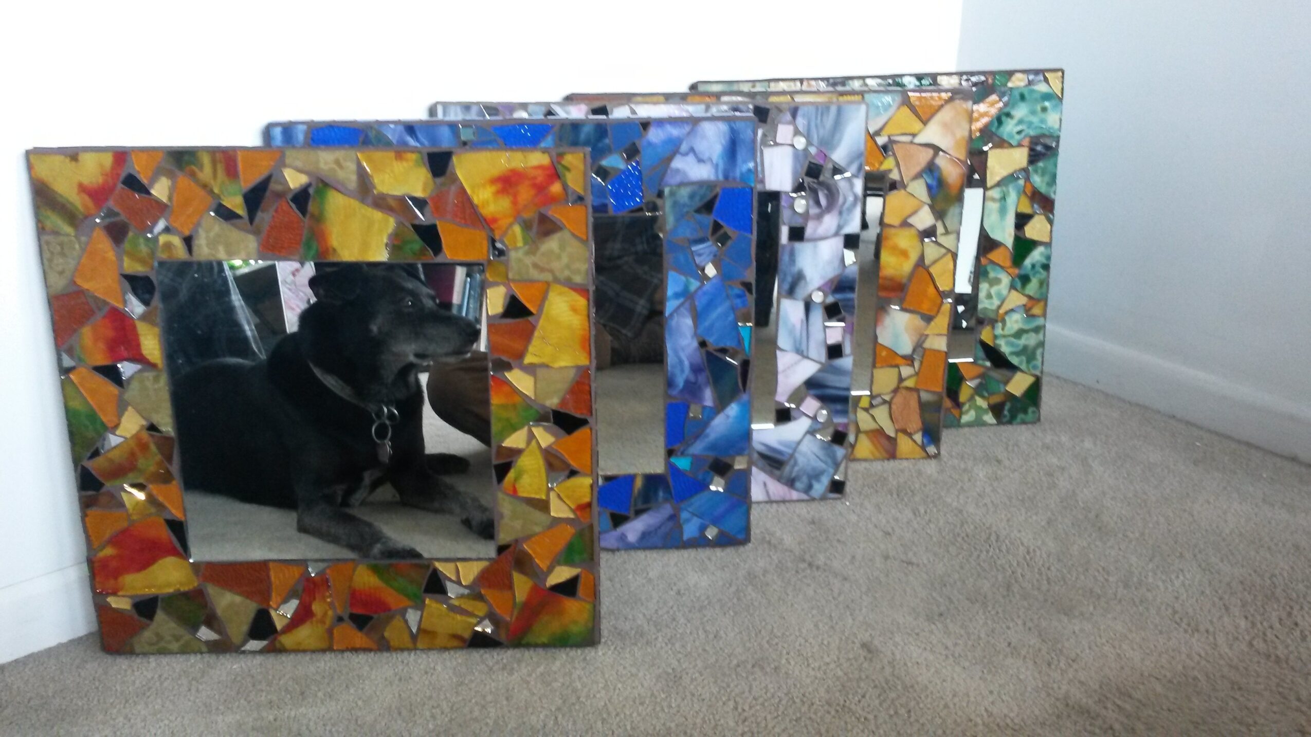 Six decorative mirrors with colorful mosaic frames leaning against a wall, one reflecting a person and a dog, on carpeted floor.