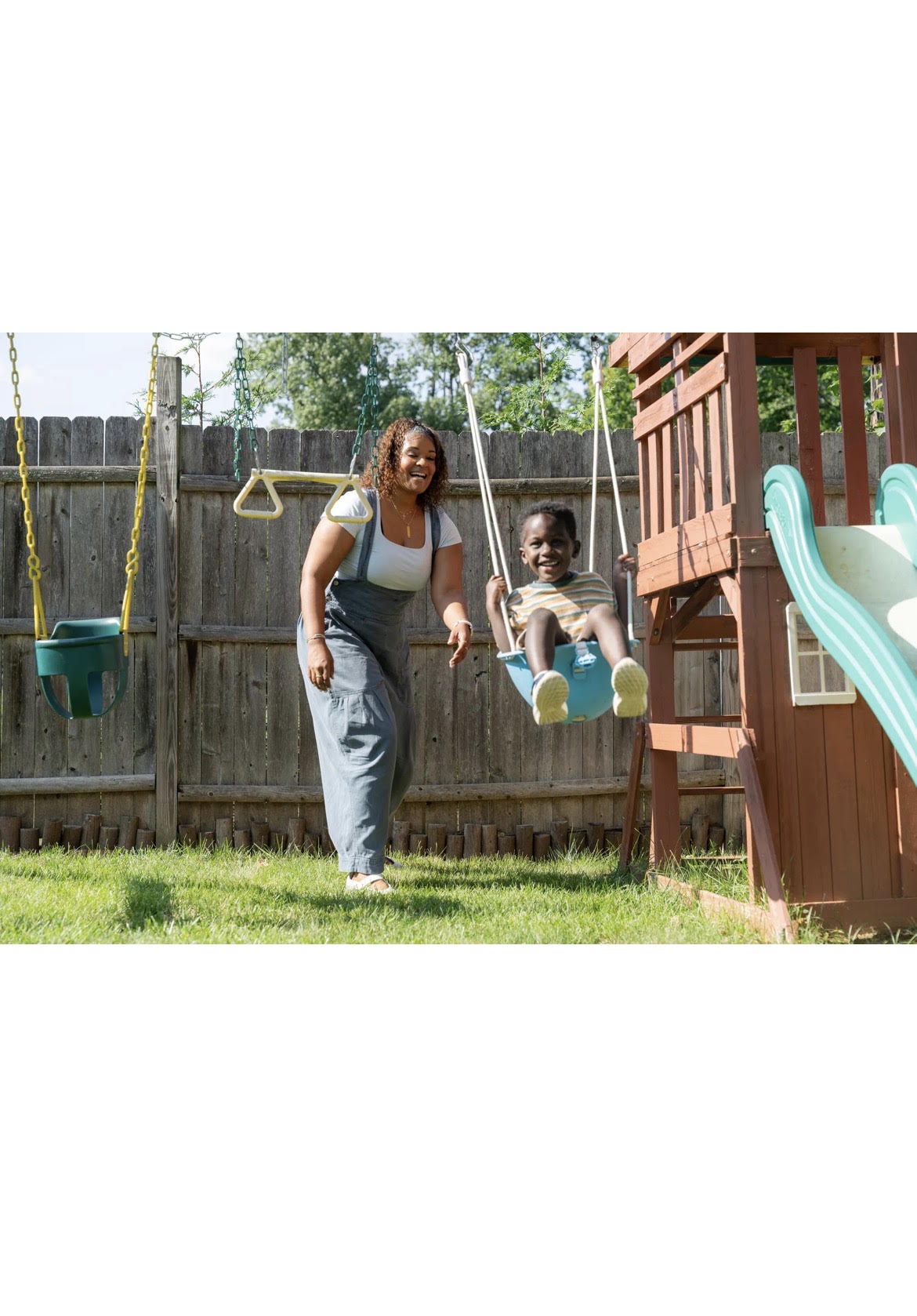 Woman pushing a child on a swing in a backyard with a wooden playset and slide, fence, and trees in background.