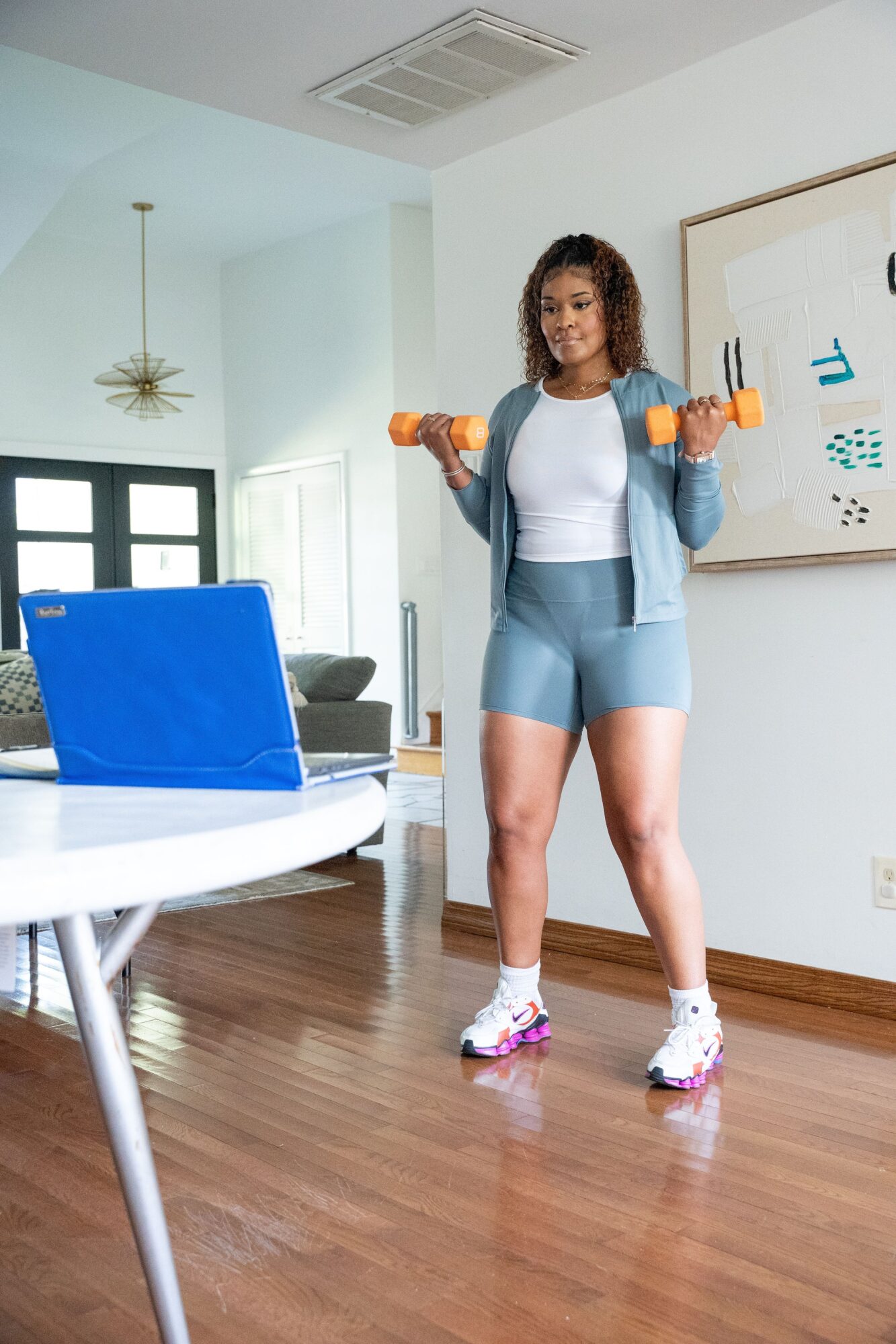 Young woman in workout clothes holding dumbbells in a bright living room with hardwood floor and white walls.
