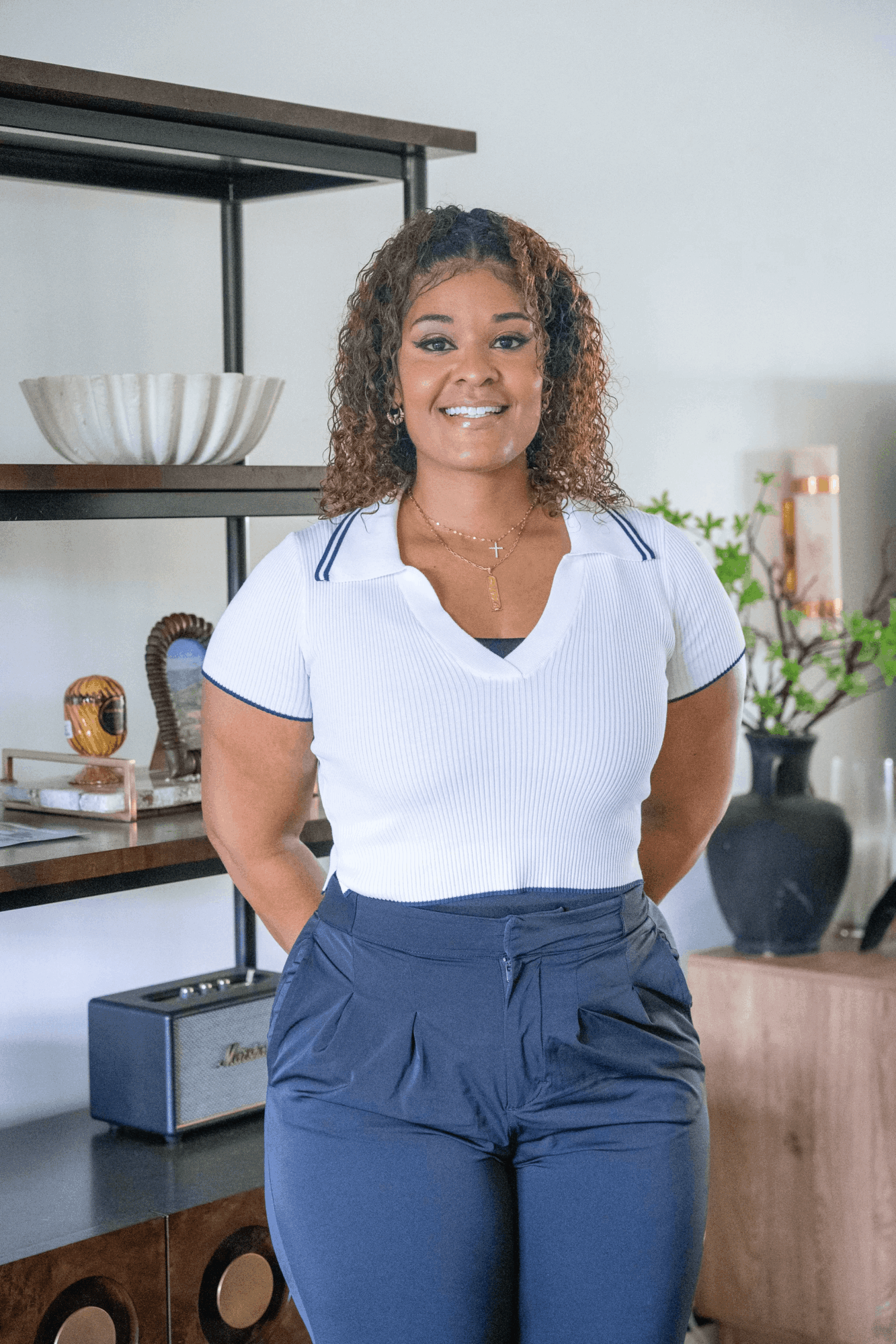Smiling woman with curly hair standing in a modern kitchen, hands behind her back, wearing a white top and dark pants.