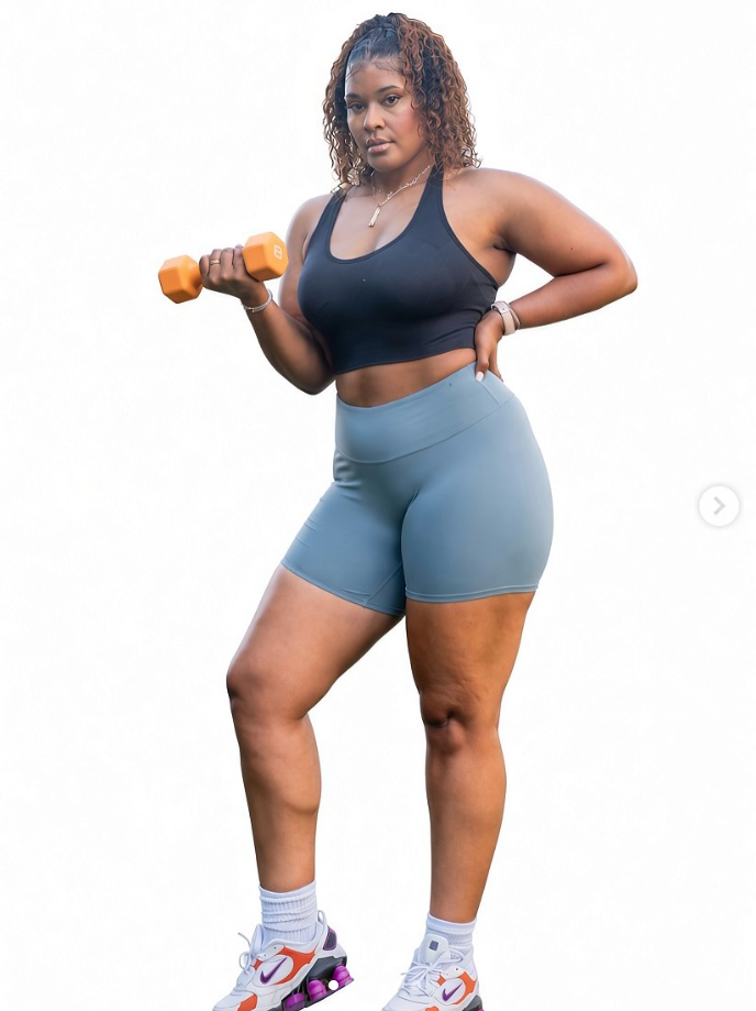 Woman in workout clothes holding a dumbbell, standing with one hand on her hip, against a white background.
