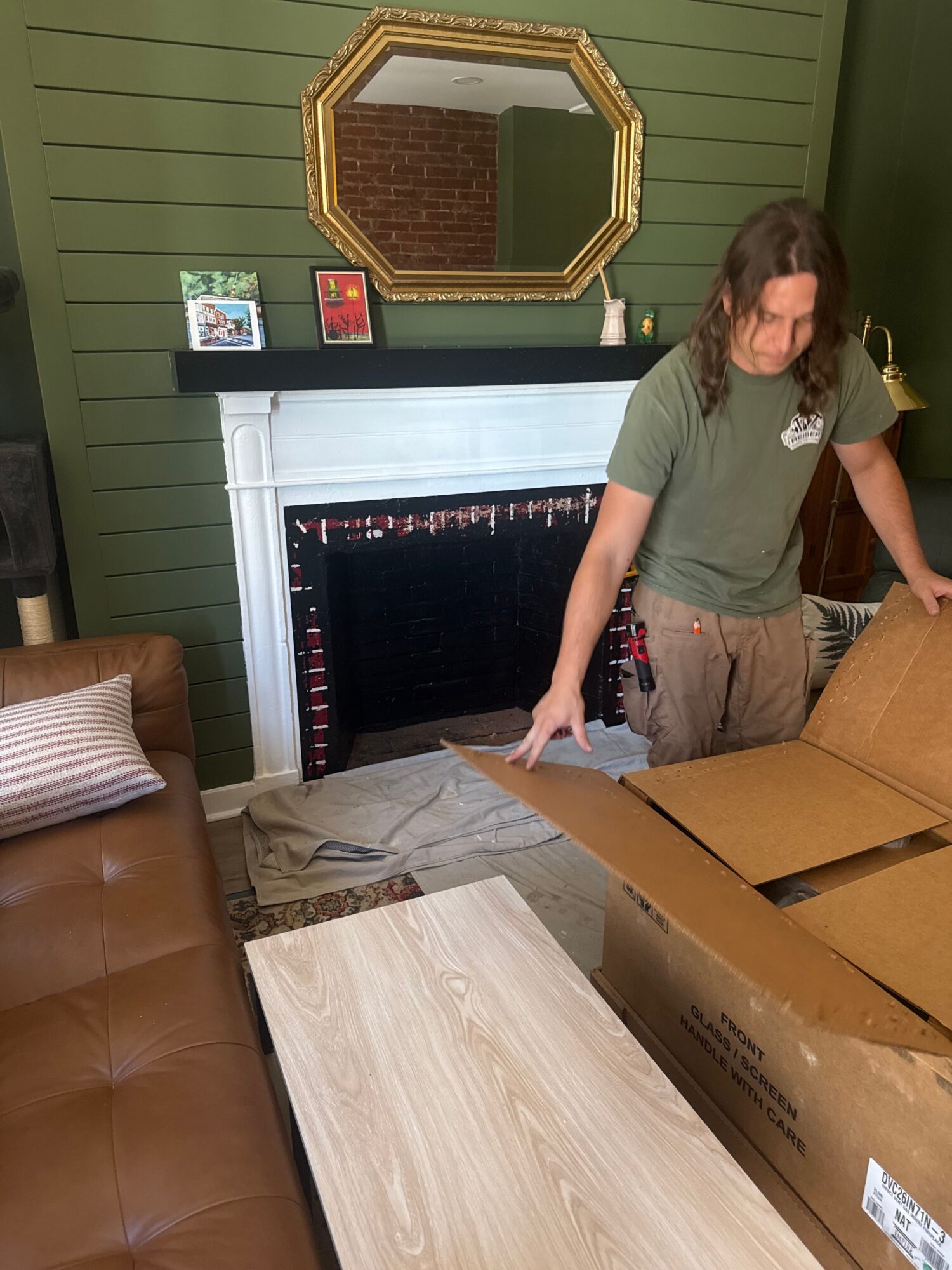 Person standing next to a table with cardboard boxes in a room with green walls and a mirror.