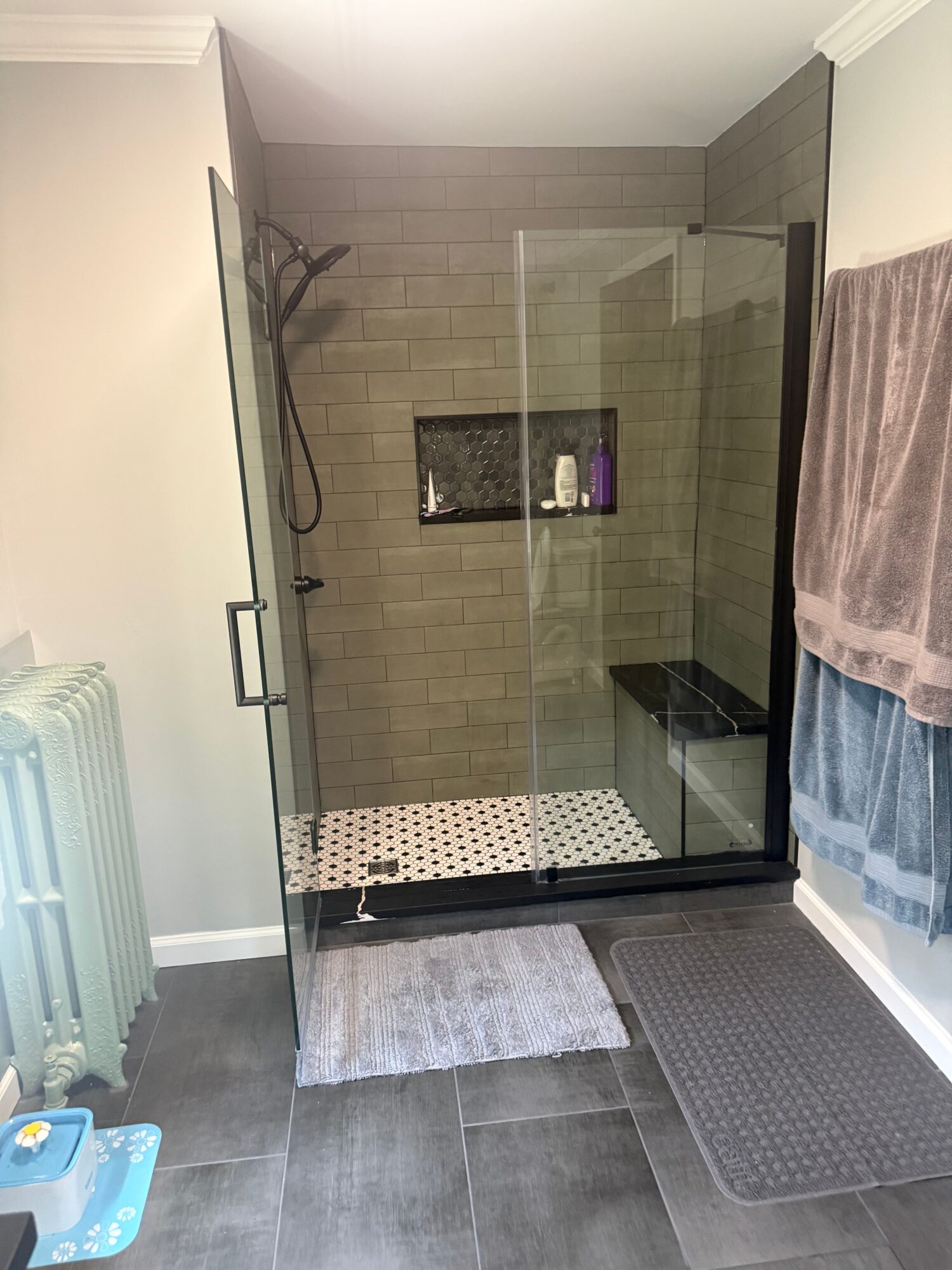Bathroom shower with glass door, tiled walls, and a small black shelf inside, viewed from outside the shower area.