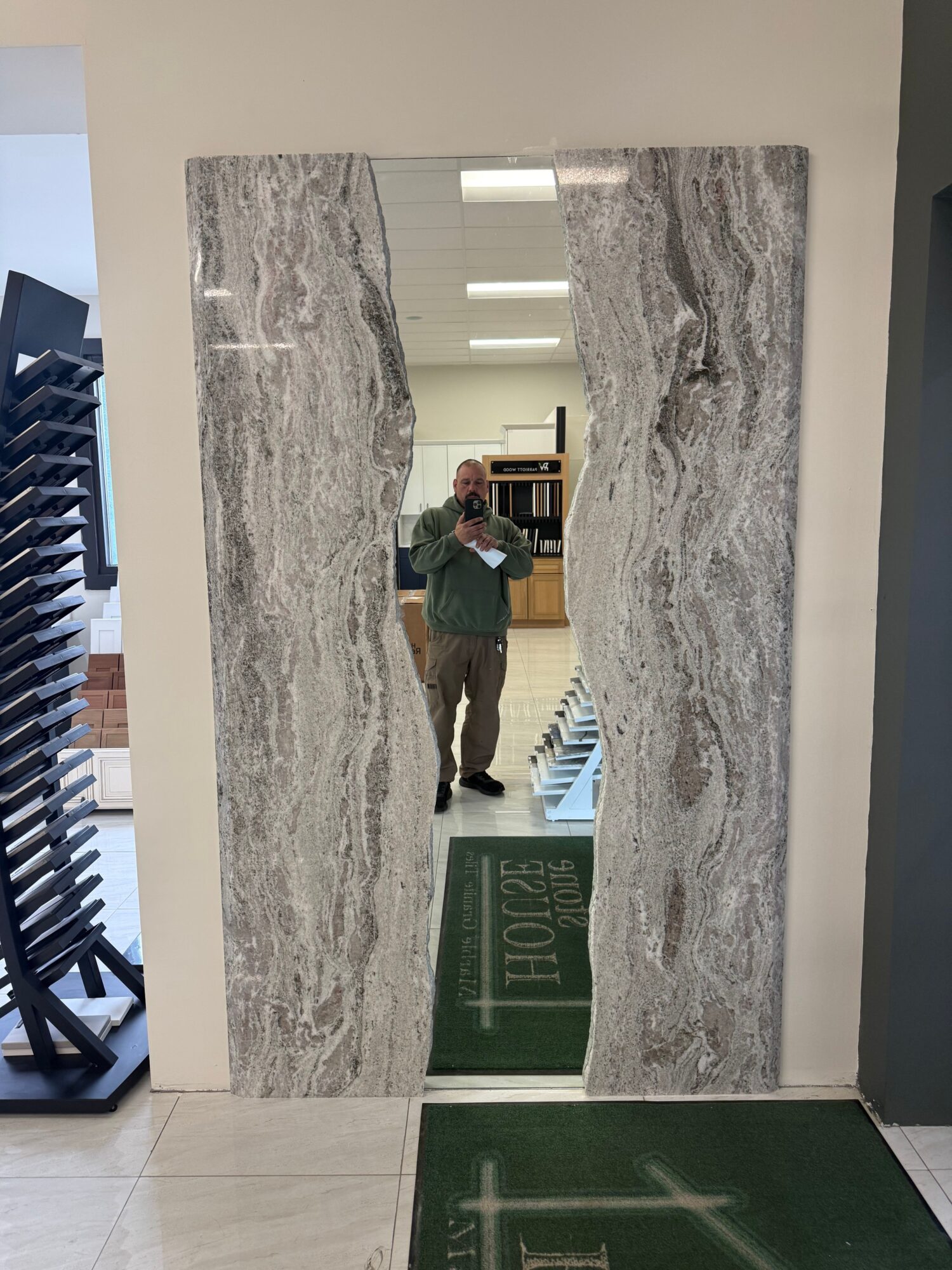 Person taking a mirror selfie in a hallway with marble walls and a green floor mat.