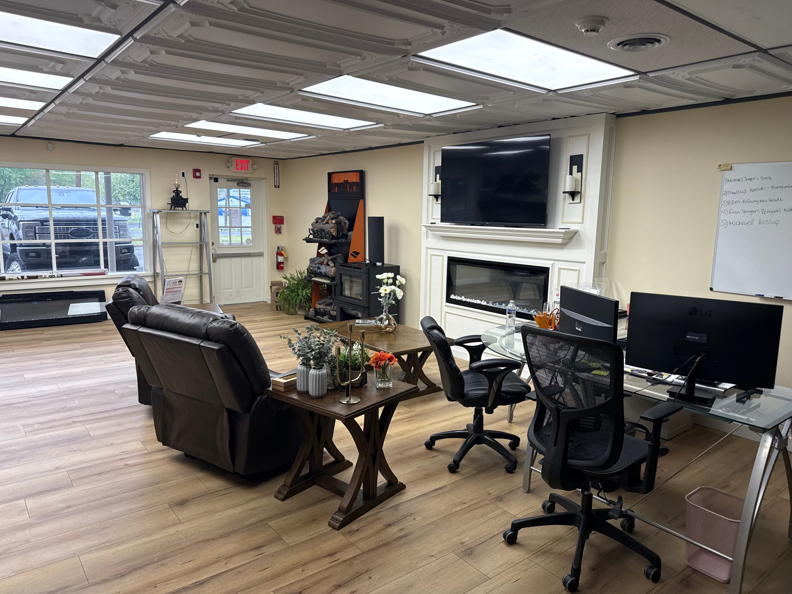 Office room with chairs, a sofa, a TV, and a whiteboard, illuminated by ceiling lights, with windows on the left side.