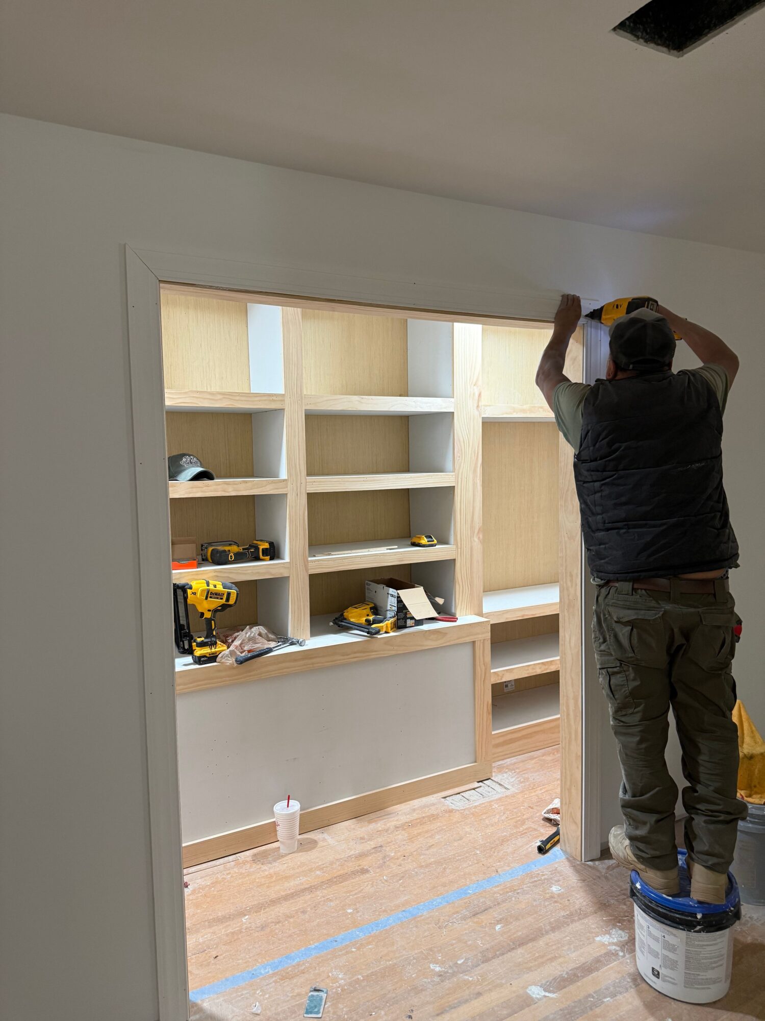 Person working on a wooden shelving unit in a room under construction, with tools and materials visible.