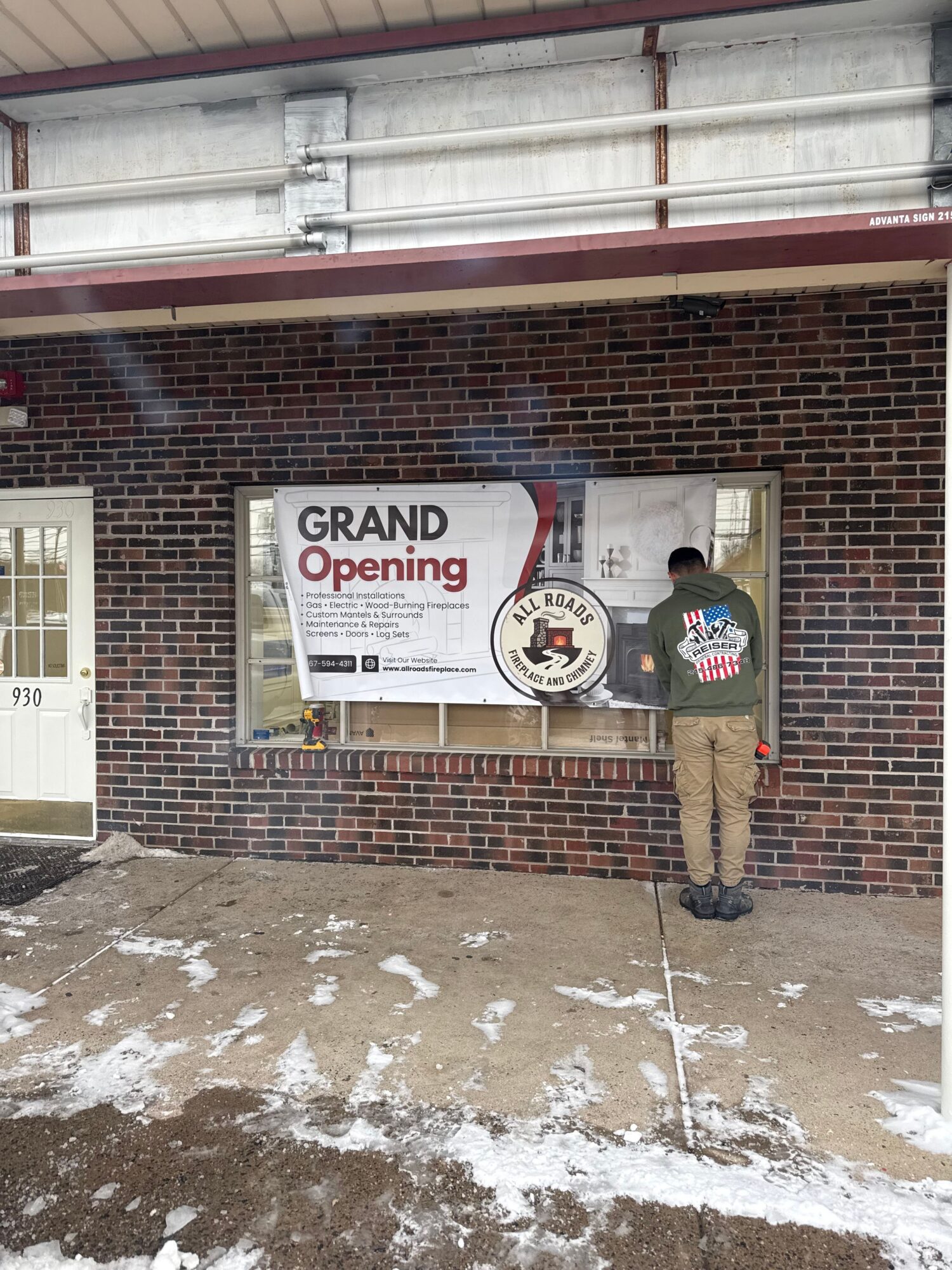A sign announces grand opening, mounted on a brick wall, with snow on the ground nearby.
